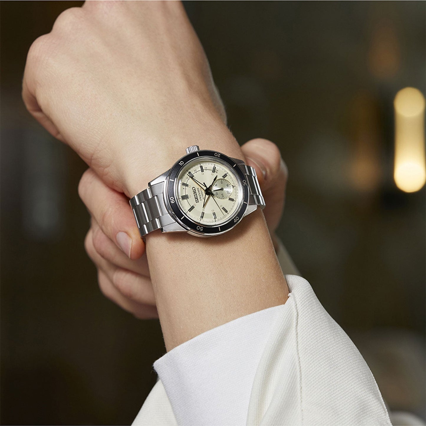 A hand wearing a Seiko Presage Style 60's Automatic With Manual Winding 40.8mm watch with a beige dial is shown. The watch, designed by Seiko, includes luminous hour markers and features an automatic movement along with a rotating bezel. The person is dressed in a white long-sleeve shirt.