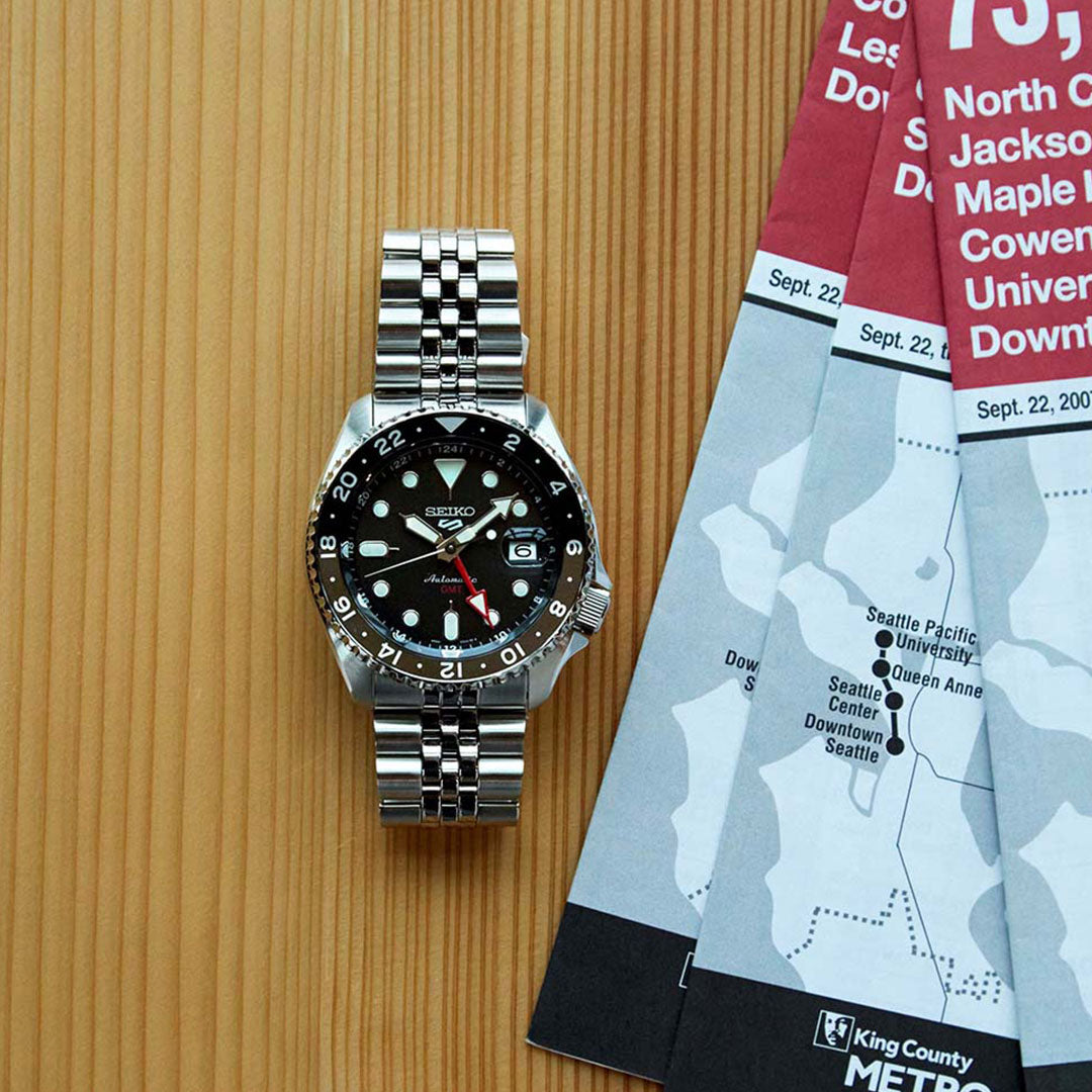 A Seiko 5 Sports SKX Series Automatic Watch, featuring a stainless steel bracelet with a black dial and red accents, rests on a wooden surface. Beside it are several folded maps showcasing destinations like the Seattle Center and University District.