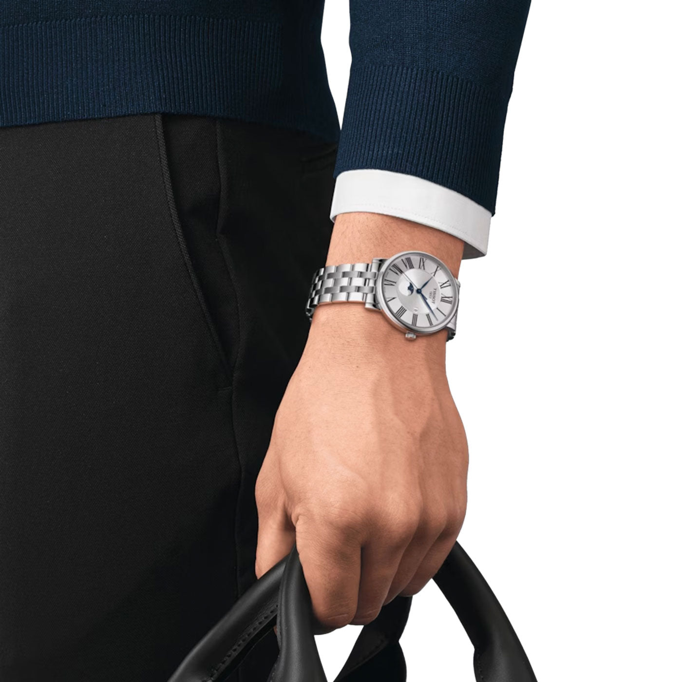 A person wearing a dark sweater holds a black leather bag. Their wrist showcases the Tissot Carson Premium Gent Moonphase Quartz 40mm Watch, featuring Swiss Quartz Movement and a white face, partially covered by a white shirt cuff. The watch's elegance is enhanced by its sapphire crystal, perfectly complementing the outfit.