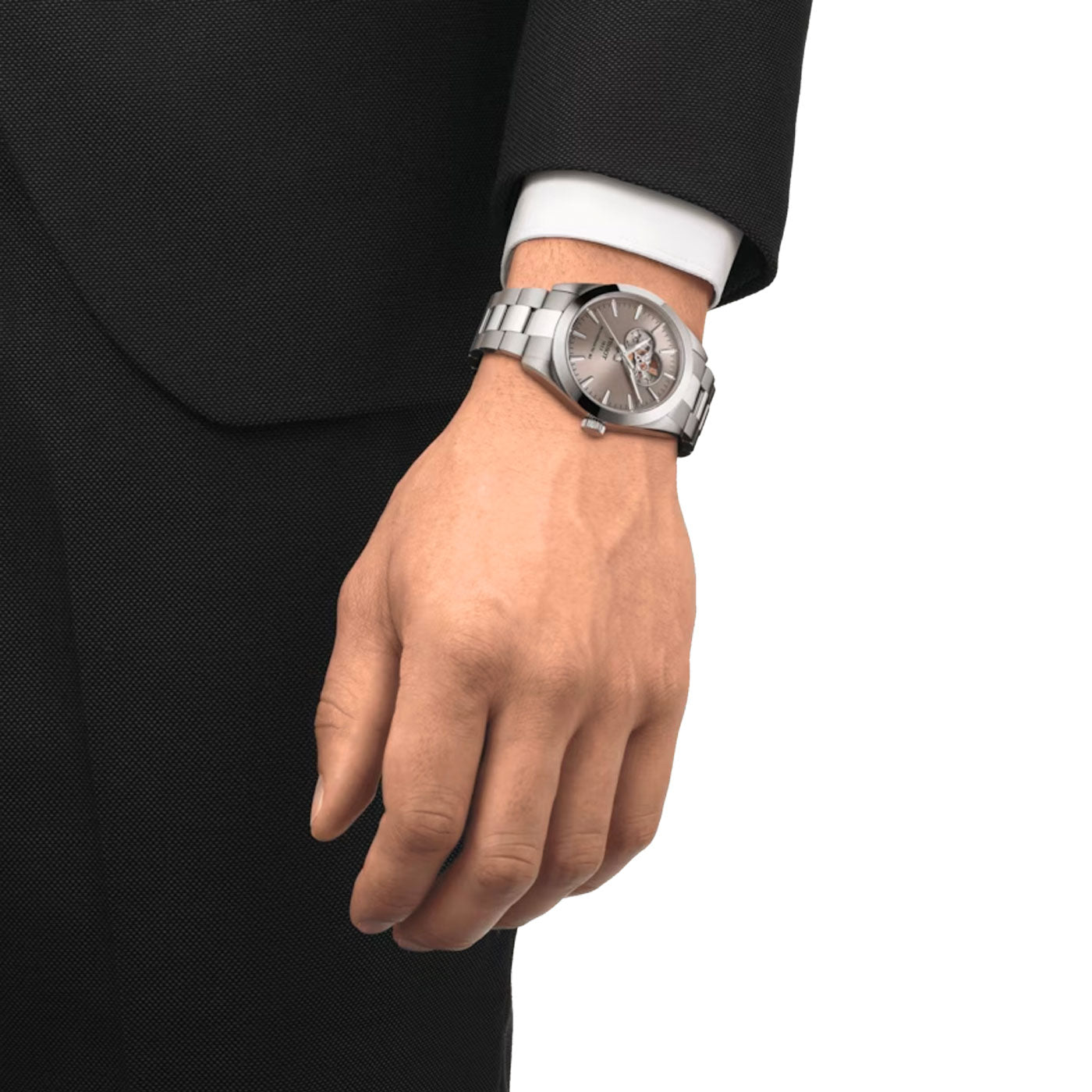 A person in a black suit with a white shirt cuff visible is wearing the Tissot Gentleman Powermatic 80 Automatic Open Heart 40mm Watch on their wrist. This silver watch showcases a sapphire crystal, has a metallic band, and features a dark face complemented by light-colored hands and markers.