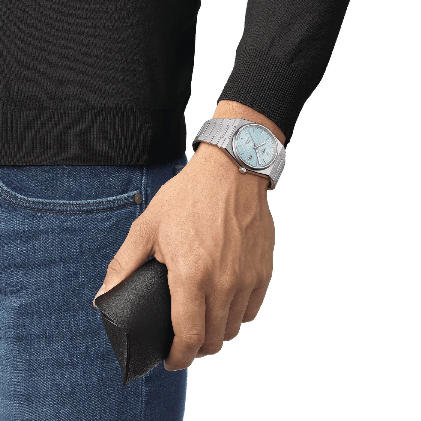 A person wearing the Tissot PRX Powermatic 80 Automatic 40mm Watch, which features a light blue face and stainless steel case, holds a black leather case in their hand. Dressed in a black sweater and blue jeans against a white background, this stylish ensemble is completed by the sophisticated watch from Tissot.