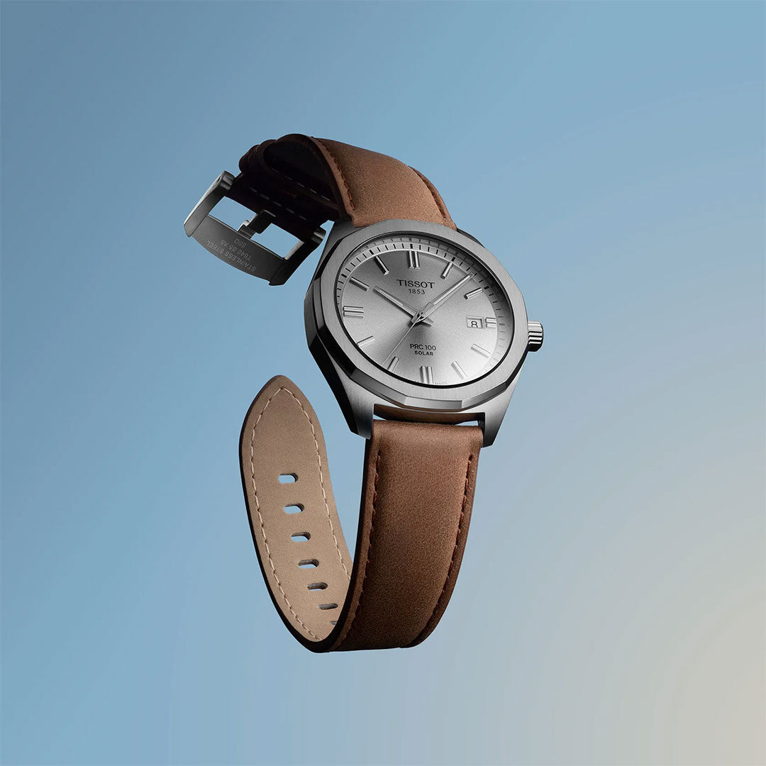 The Tissot PRC 100 Solar Quartz 39mm Watch by Tissot, featuring a silver case and light brown leather strap, floats against a gradient blue background. Its dial shows the time and date with minimalist hour markers and hands.