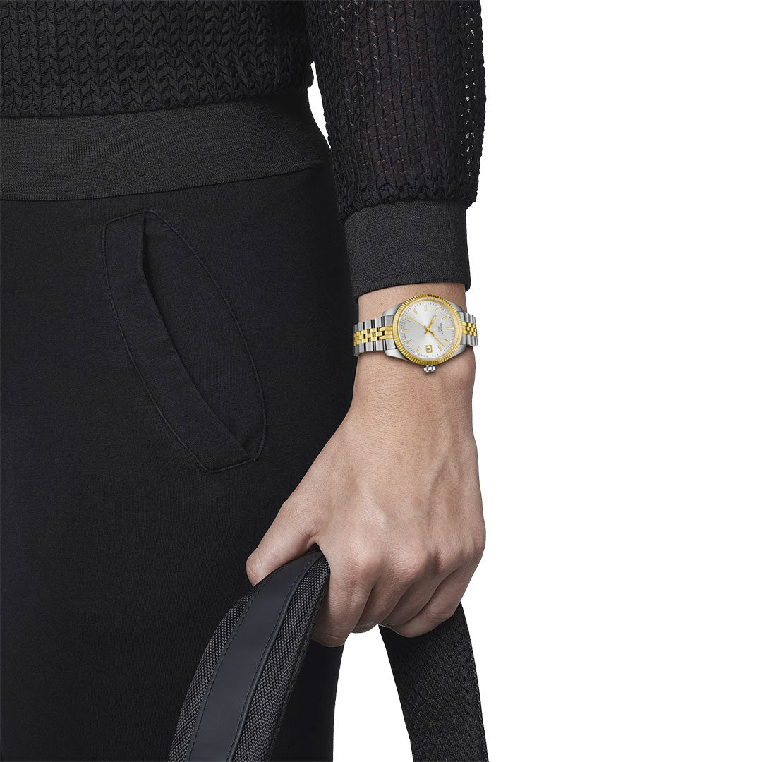A person in black attire holds a black textured strap, displaying the Tissot Ballade Quartz 34mm Watch with a two-tone silver and gold fluted bezel. Only the lower torso, arm, and hand are visible against a white background.