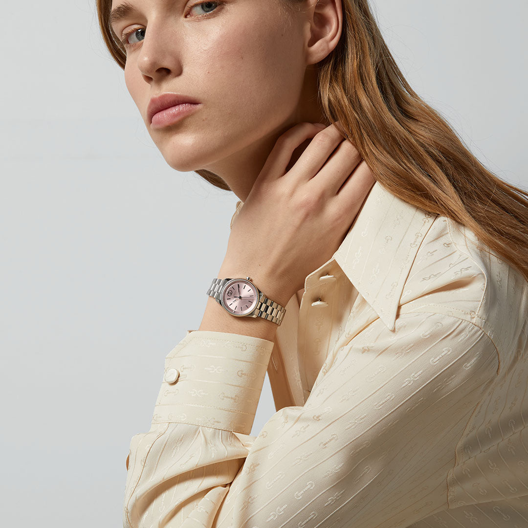 A woman with long light brown hair, in a cream button-up shirt, poses with her hand near her neck, showcasing the GUCCI G-Timeless Quartz 29mm Watch with a pink face and stainless steel bracelet.