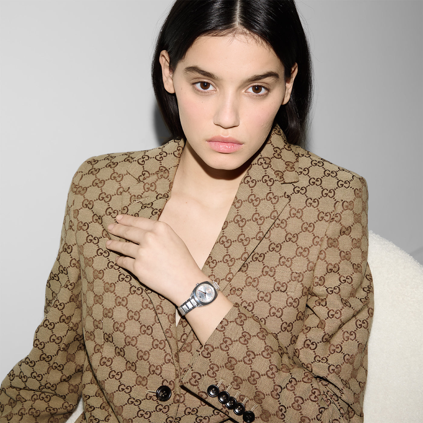 A person with long dark hair wears a brown patterned blazer and a Gucci Interlocking G Quartz 29mm Watch by GUCCI, sitting against a light background. Their hand rests near their chest.