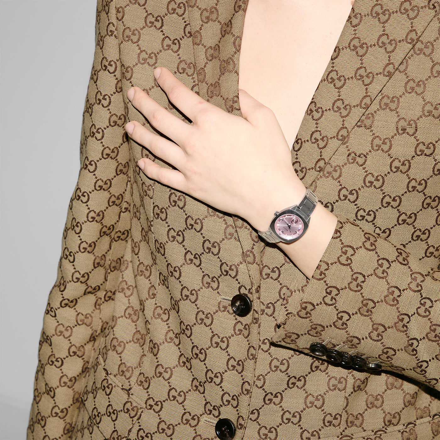 A person wearing a patterned brown suit with a matching jacket gently holds the lapel while showcasing the stylish Gucci Interlocking G Quartz 29mm Watch, featuring a silver sunbrushed dial. The suit's distinctive repeating design beautifully complements the elegance of their GUCCI timepiece.