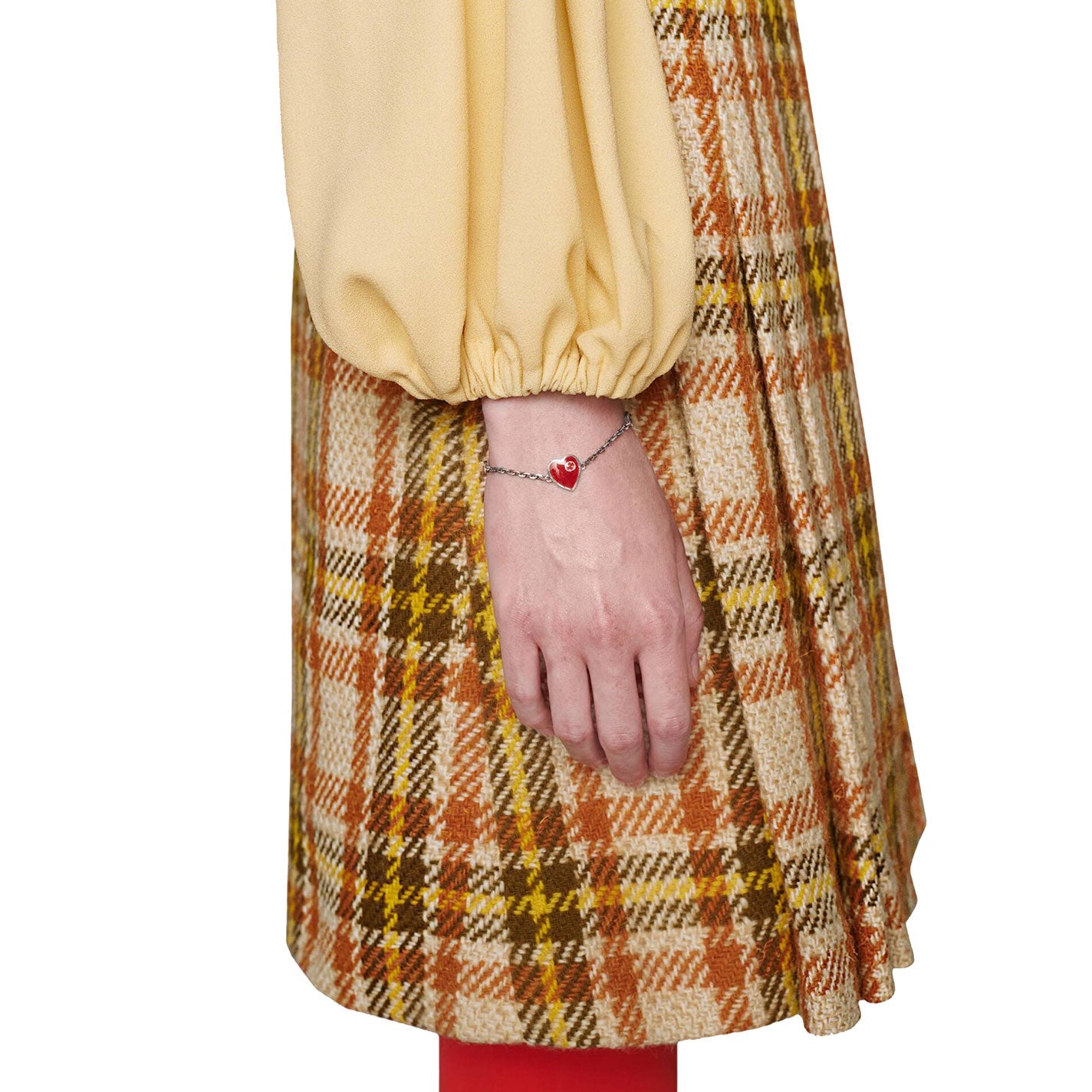 A person wearing a long-sleeve yellow top with a plaid skirt in brown, orange, and yellow tones is holding a GUCCI Fine Jewellery Interlocking G Sterling Silver Station Bracelet with a red enamel heart pendant. Red tights can be seen peeking out from under the skirt.