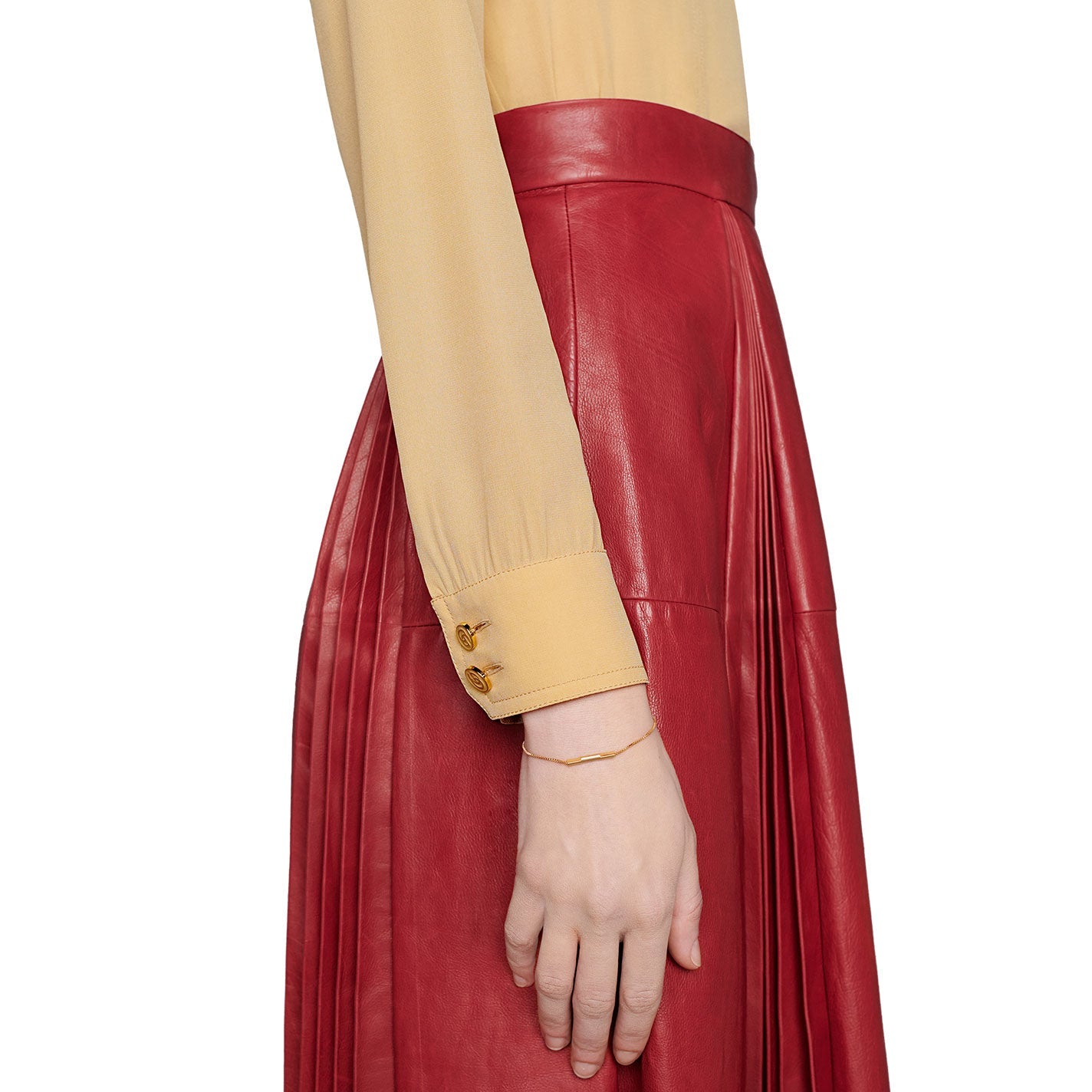 A person wearing a beige long-sleeve shirt with buttoned cuffs and a pleated red leather skirt, adorned with the Gucci Link to Love 18K Yellow Gold Chain Bracelet With "Gucci" Bar from GUCCI Fine Jewellery. The side view highlights the sleeve details, the texture of the skirt, and the bracelet glinting as the hand rests elegantly beside it.