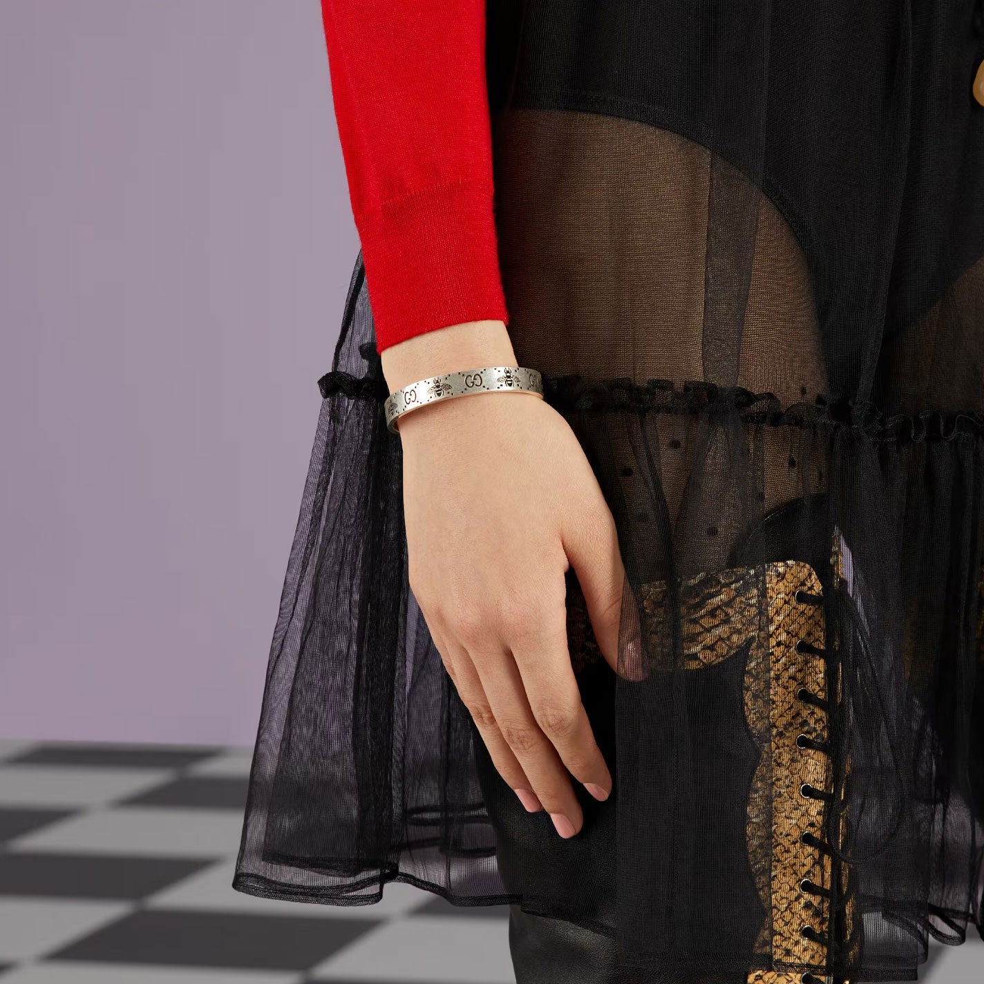 A person wearing a red sleeve is showcasing the Gucci GG And Bee Engraved Sterling Silver Cuff Bracelet from GUCCI Fine Jewellery, which features a subtle design. The skirt is sheer black with frill details, revealing patterned leggings underneath, set against a backdrop of a checkered floor and a plain wall.