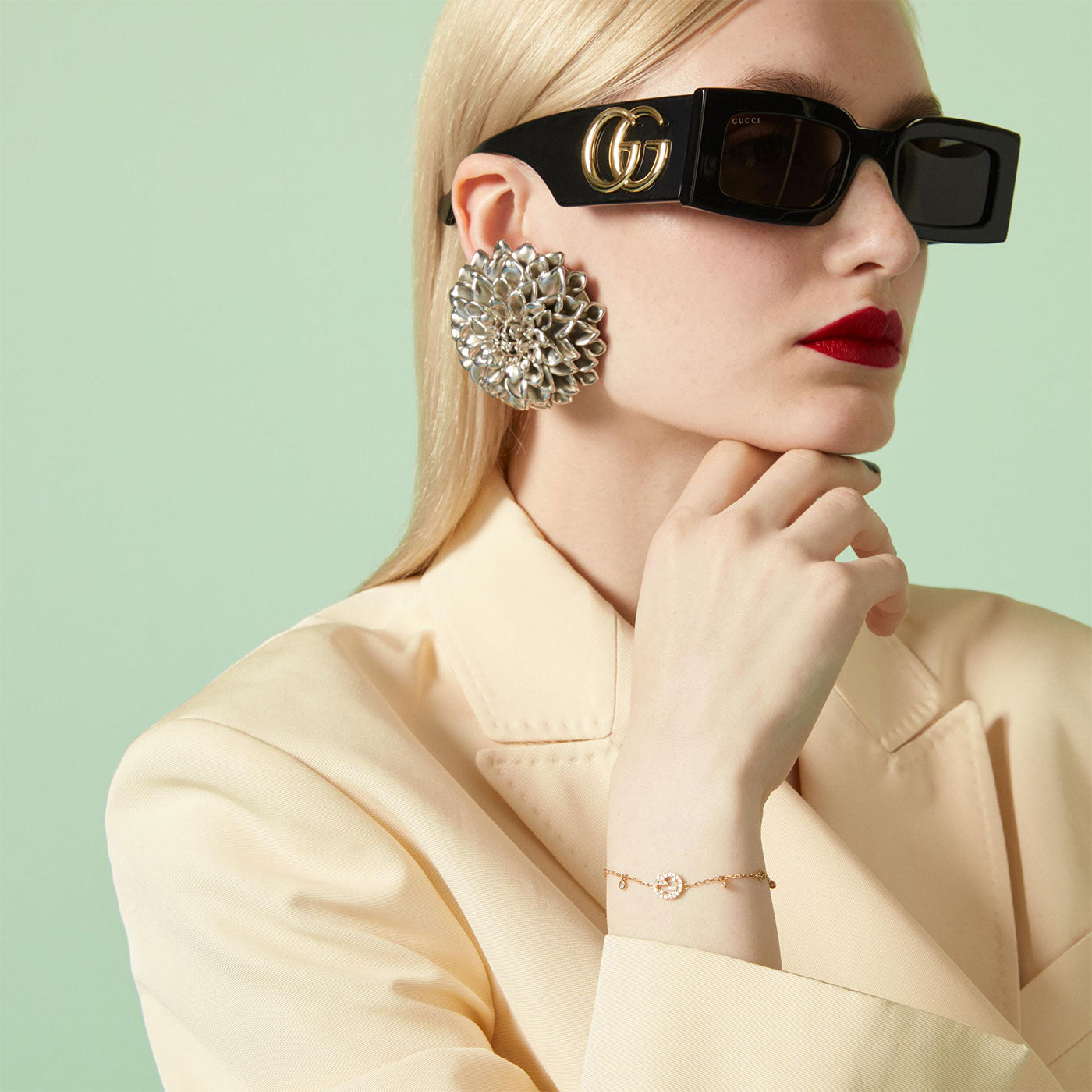 A person with blonde hair wears large black sunglasses, a cream blazer, a striking silver earring, and the Gucci Interlocking G 18K Yellow Gold Diamond Bracelet by GUCCI Fine Jewellery. The background is light green, and the person has a thoughtful pose with red lipstick.