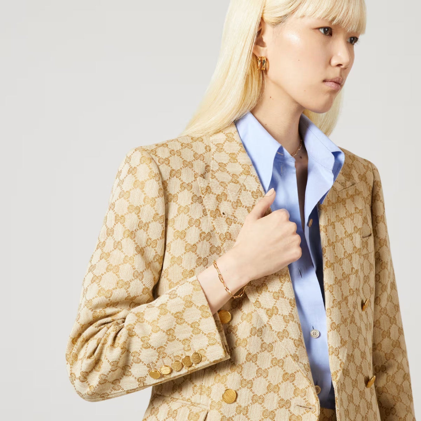 A person with long blonde hair wears a light blue shirt and a beige jacket with a patterned design. A touch of sophistication is added by the Gucci Link to Love 18K Yellow Gold Chain Bracelet from GUCCI Fine Jewellery, peeking just above the cuffs, as their hand rests near the jacket's label against a plain, light background.