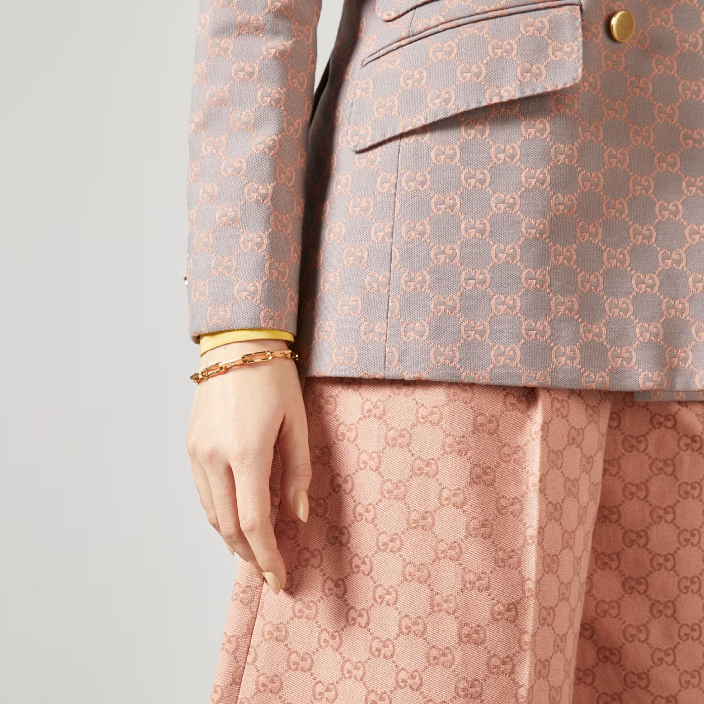 A person wearing a stylish outfit with a gray Gucci blazer and pink Gucci pants, featuring the signature GG pattern, casually rests their hand by their side, showcasing a Gucci Link to Love 18K Yellow Gold Wide Chain Bracelet from GUCCI Fine Jewellery on their wrist.