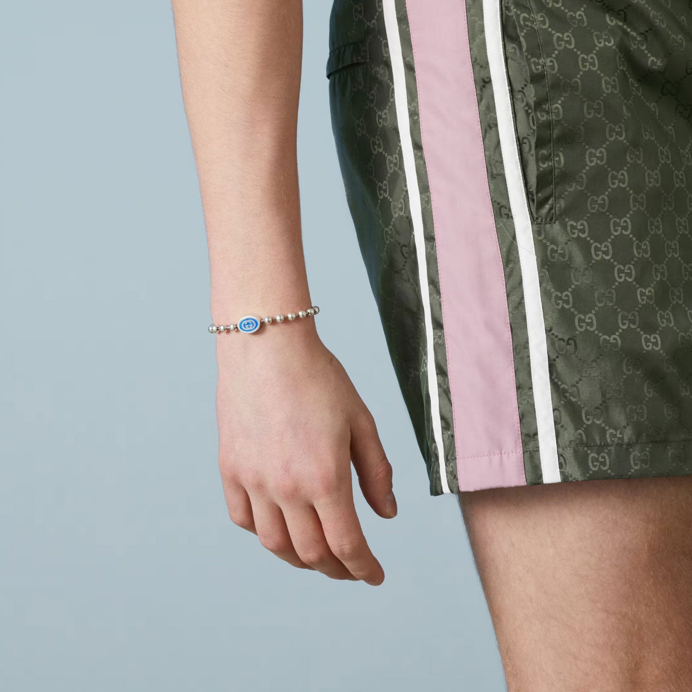 A person wearing green shorts with pink and white stripes stands against a light blue background. On their wrist, they sport the Gucci Interlocking G Boule Sterling Silver Bead Bracelet from GUCCI Fine Jewellery, adorned with a blue enamel charm.