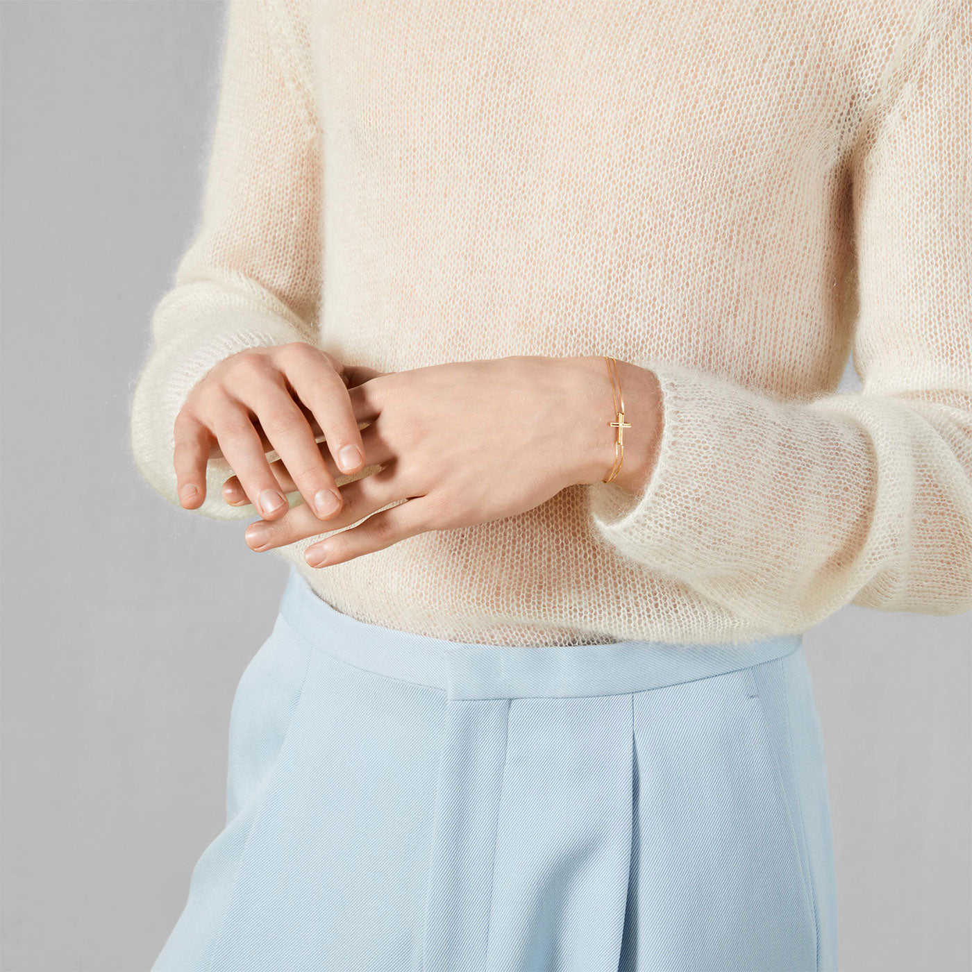 A person dressed in a cream sweater and light blue pants stands with their hands together, adorned with the GUCCI Fine Jewellery Gucci Link to Love Cross Charm 18K Yellow Gold Bracelet. The background is a neutral gray.