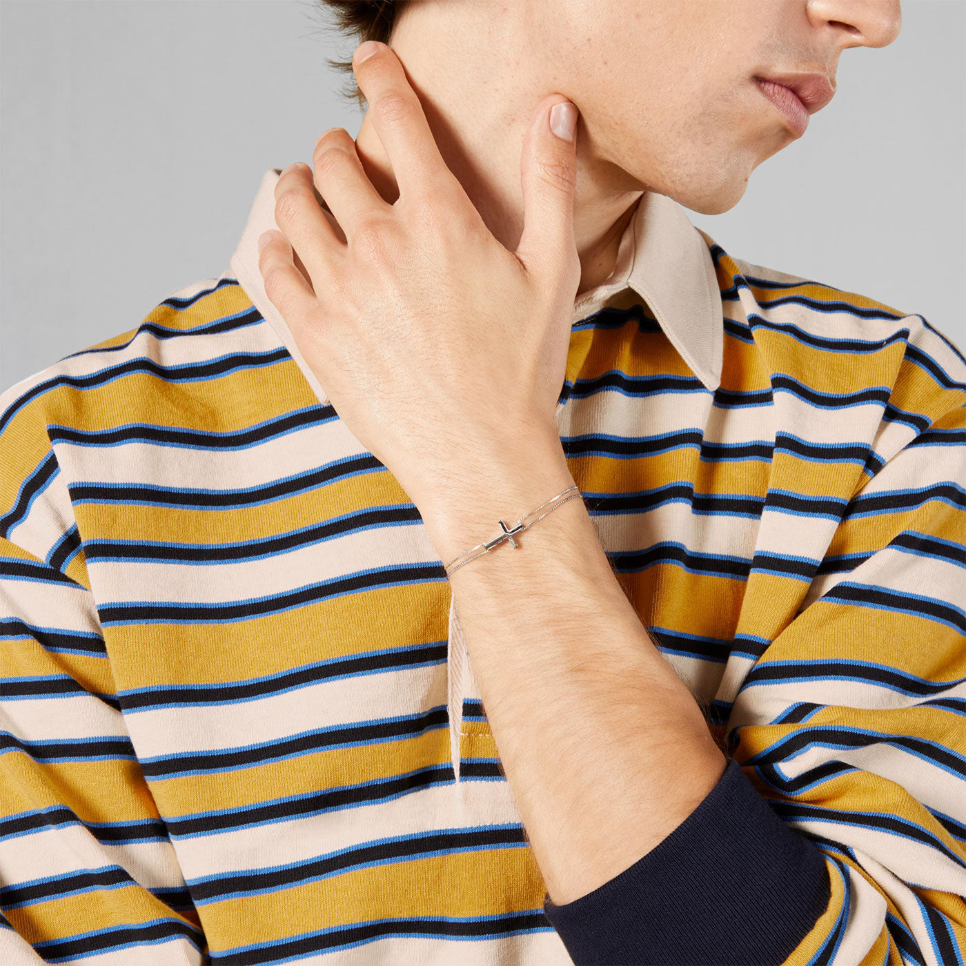 A person wearing a striped yellow and navy shirt holds their hand near their shoulder, showcasing the Gucci Link to Love Cross Charm 18K White Gold Bracelet by GUCCI Fine Jewellery. The plain gray background accentuates the elegance of this exquisite piece.