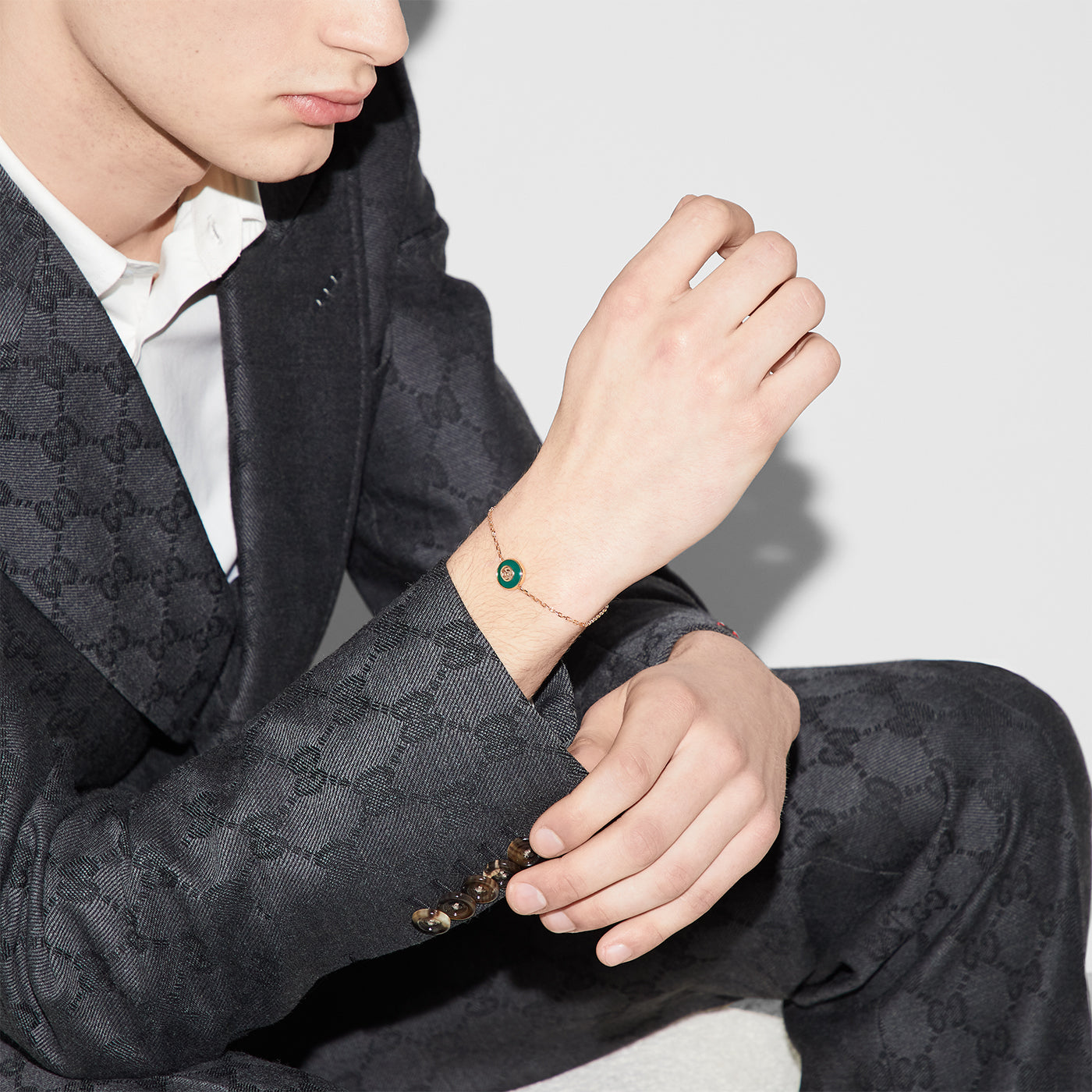 A person dressed in a dark textured suit sits against a light background, wearing the Gucci Interlocking G 18K Rose Gold Chain Bracelet With Green Agate. Their head is slightly turned, focusing on their wrist adorned with this piece from GUCCI Fine Jewellery.