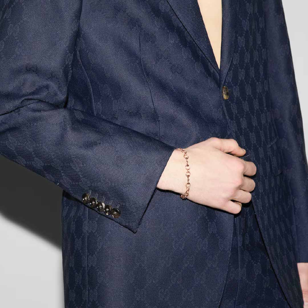 A person in a dark patterned suit, partially unbuttoned, rests their hand on their stomach. Their wrist sports the Gucci Horsebit 18K Rose Gold Diamond Station Bracelet by GUCCI Fine Jewellery.
