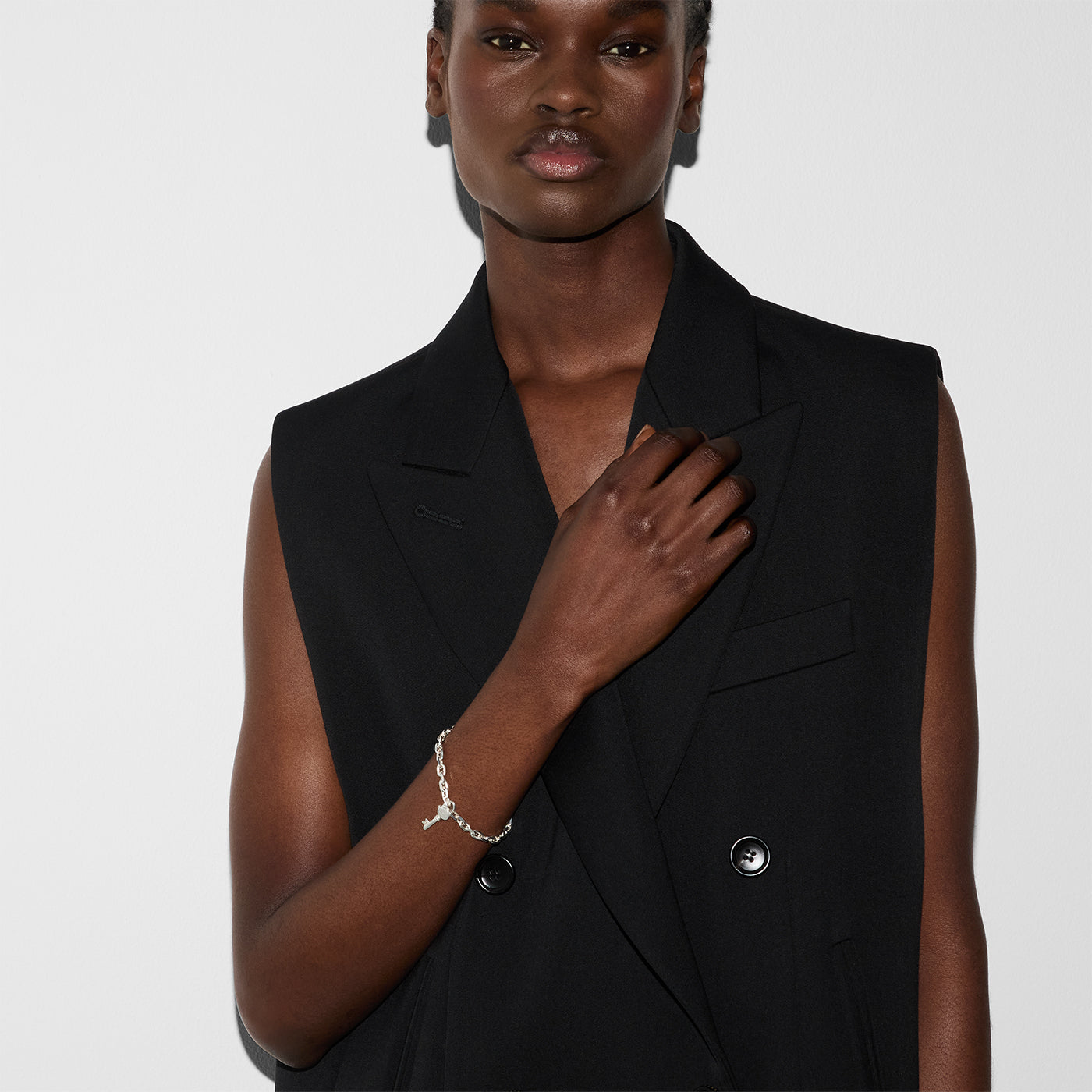 A person wearing a sleeveless, black double-breasted vest poses against a plain white background. They touch the lapel with one hand while wearing a delicate Gucci Trademark Sterling Silver Chain Bracelet With Key from GUCCI Fine Jewellery.