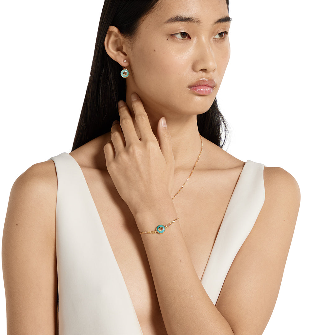 A woman with long dark hair in a sleeveless white V-neck top wears GUCCI Fine Jewellery's Interlocking 18K Yellow Gold Diamond Bracelet with Amazonite, pairing it with matching earrings and necklace as she gazes to the side neutrally.