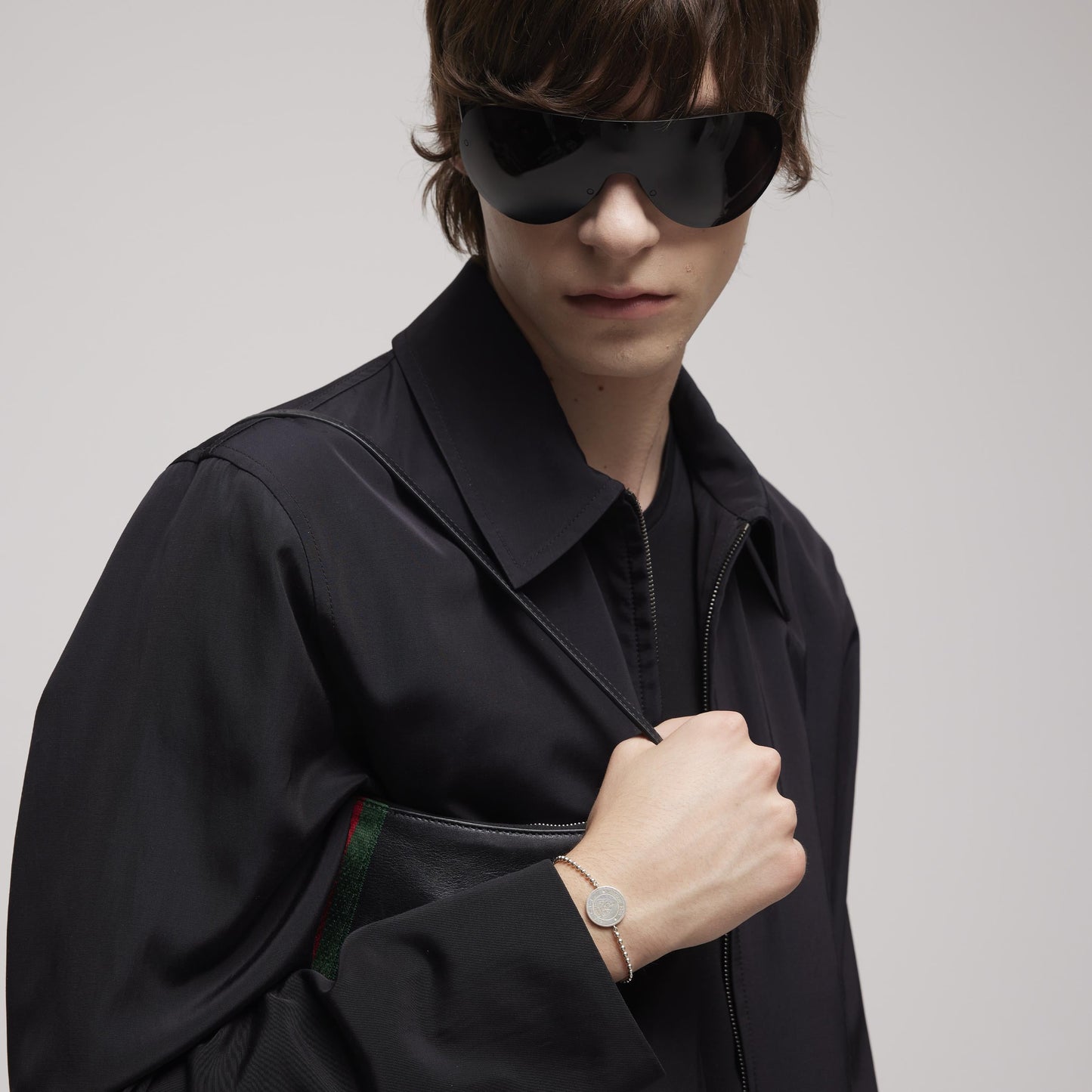 A person wearing large black sunglasses and a dark jacket holds a black clutch under their arm, accessorized with the GUCCI Fine Jewellery Gucci Crest Sterling Silver Boule Bracelet that complements their short brown hair and modern look.