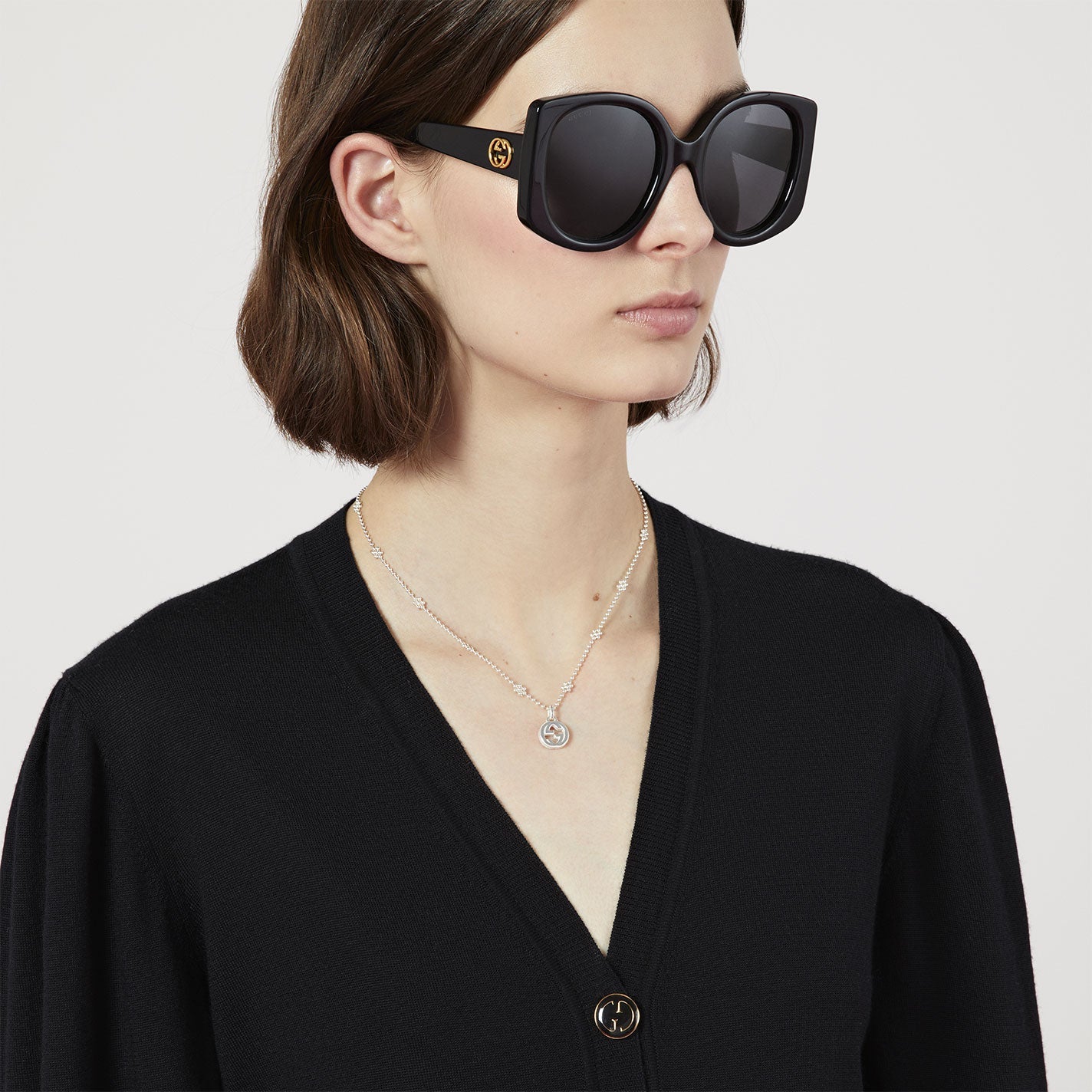 A person with shoulder-length brown hair is wearing large, square black sunglasses and a black cardigan. The subtle elegance of the Gucci Interlocking G Sterling Silver Necklace Pendant from GUCCI Fine Jewellery graces their neck. The backdrop is a simple white canvas.