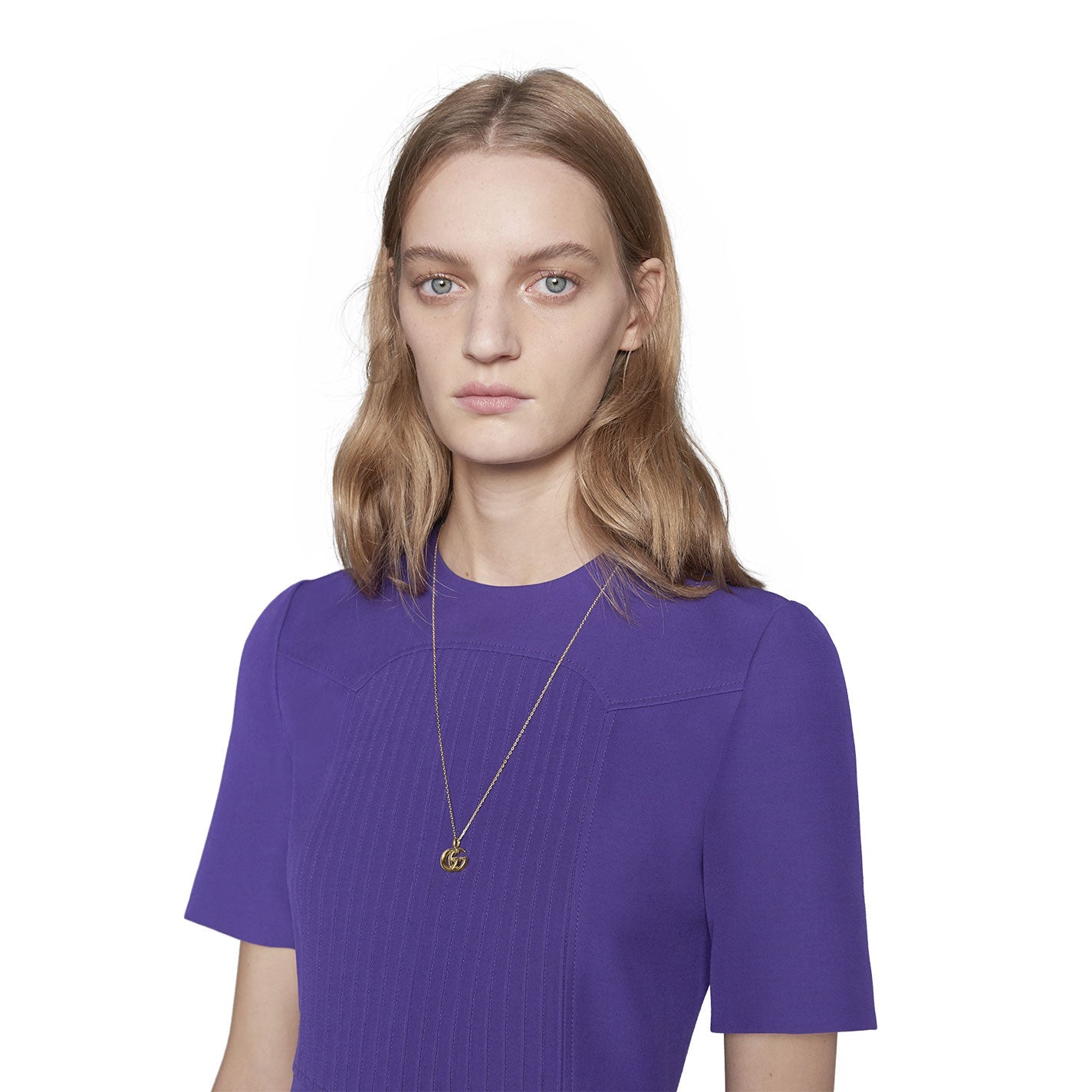 A person with long, light brown hair wears a purple crew-neck top paired with the Gucci GG Running 18K Yellow Gold Necklace Pendant. This elegant piece from GUCCI Fine Jewellery features a circular pendant that exudes timeless style against a plain white background.