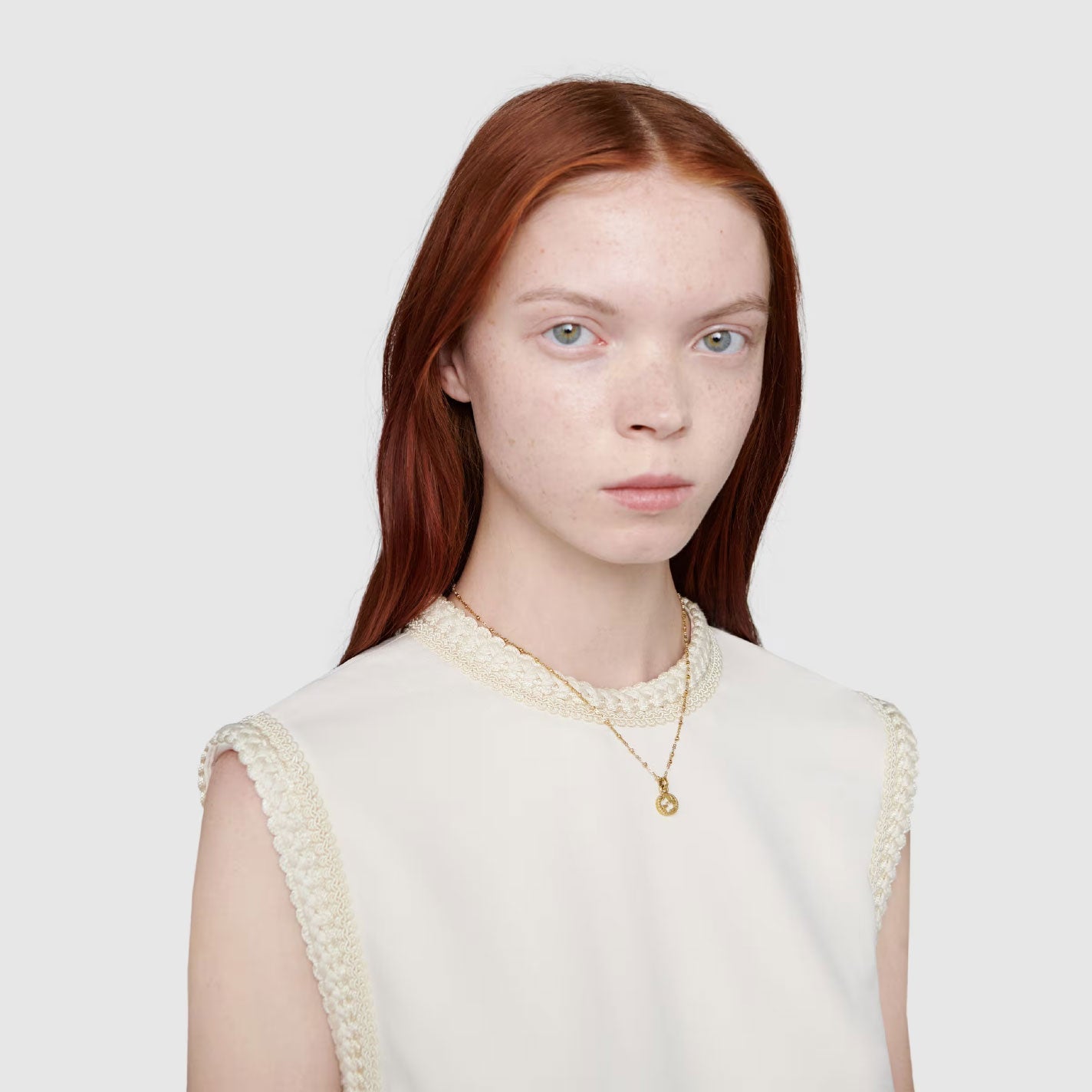 A person with long red hair wearing a sleeveless white top and a Gucci Interlocking G 18K Yellow Gold necklace pendant from GUCCI Fine Jewellery stands against a plain gray background.