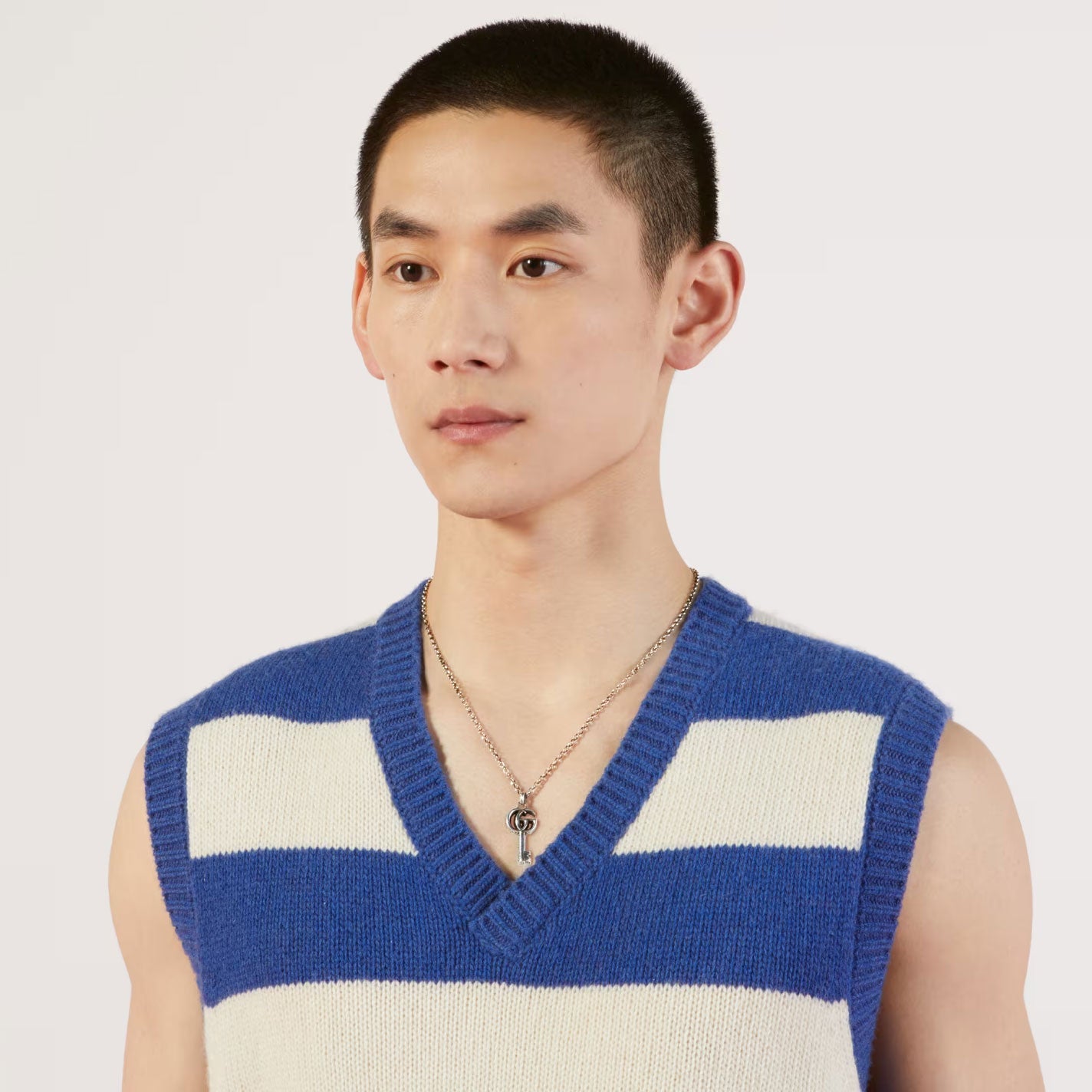 A person with short hair is wearing a blue and white striped sleeveless sweater and a GUCCI Fine Jewellery necklace featuring the Gucci Double G Key sterling silver pendant, standing against a plain white background.