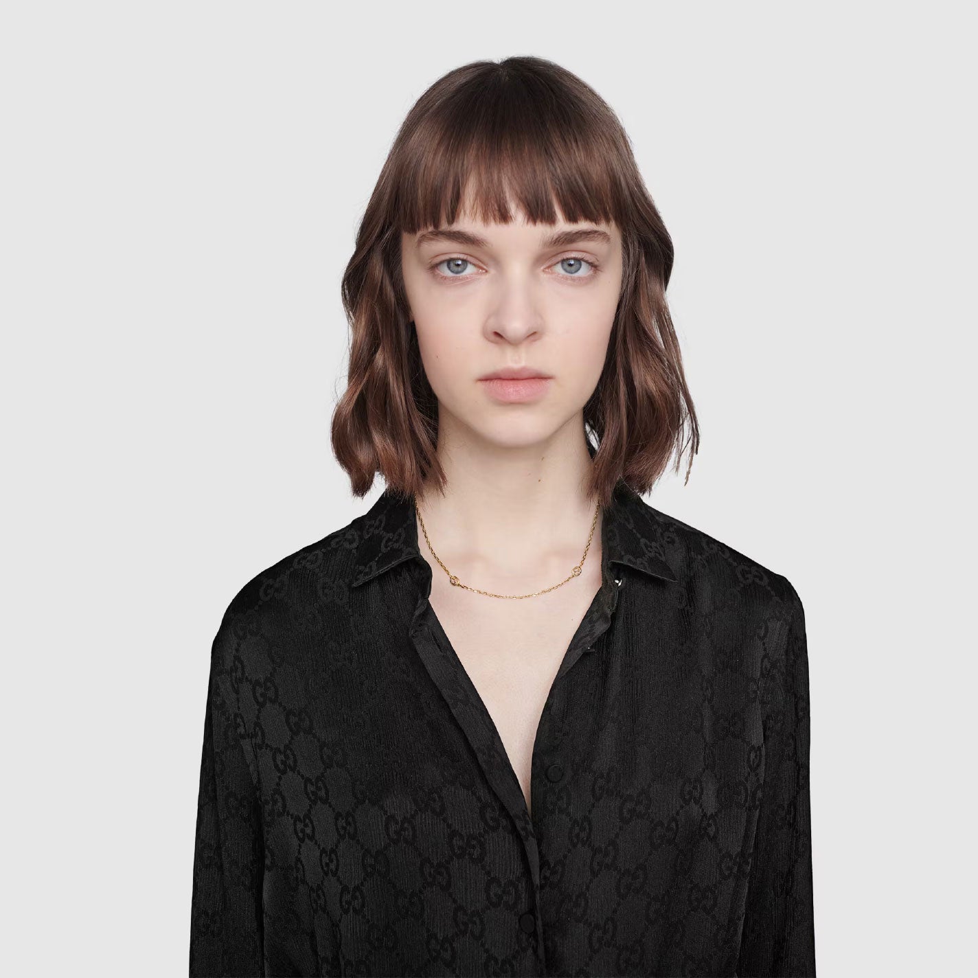 A person with shoulder-length brown hair poses against a light gray background, wearing a black Gucci shirt with a subtle pattern. Their expression is neutral, and they are accessorized with the GUCCI Fine Jewellery Interlocking G 18K Yellow Gold Station Necklace.