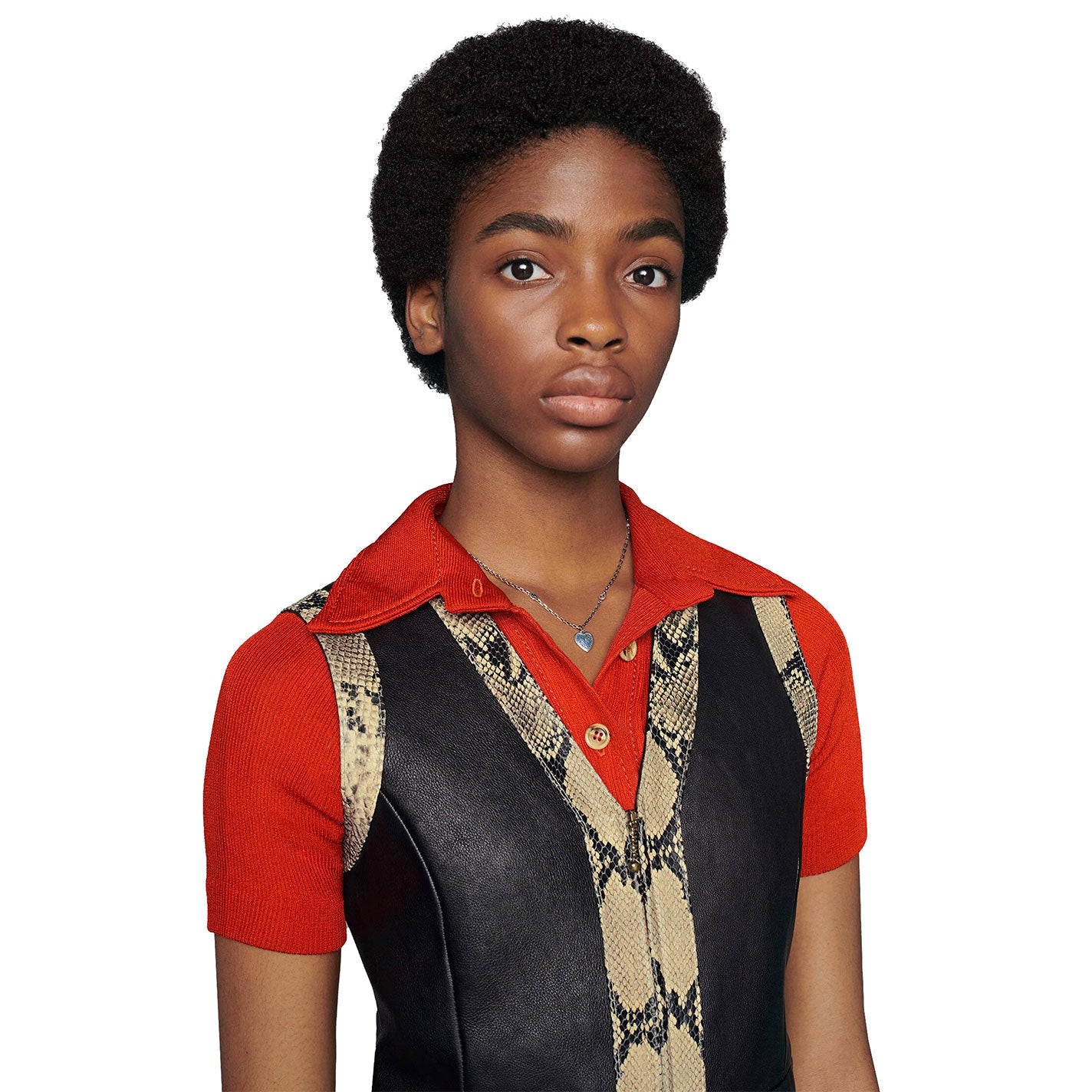 A person with short, curly hair, dressed in a red collared shirt and a vest showcasing a snakeskin pattern, stands against a plain white background. Their look is subtly highlighted by the Gucci Interlocking G Sterling Steel Necklace with Blue Enamel Heart Pendant from GUCCI Fine Jewellery as they maintain a neutral expression while gazing directly at the camera.