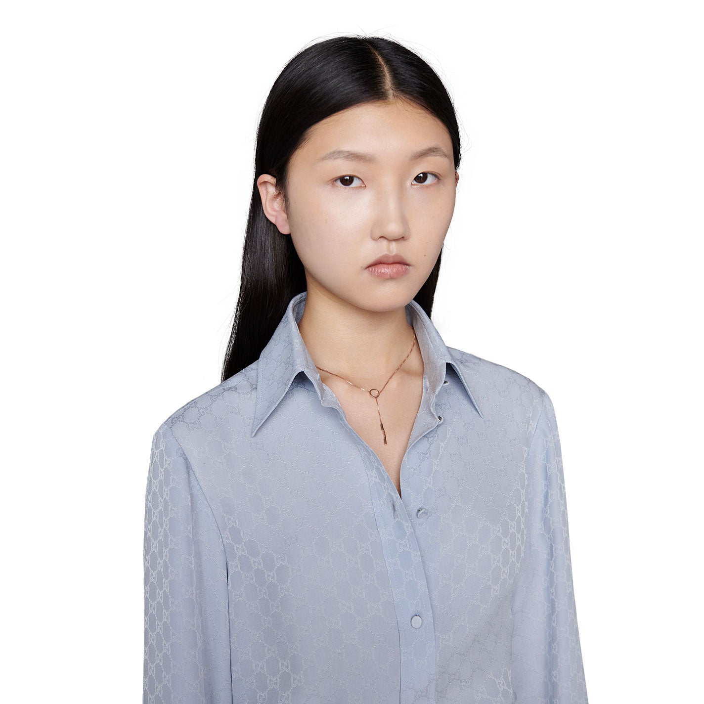 A person with long black hair is wearing a light blue blouse with a subtle pattern. They have a neutral expression and are adorned with the Gucci Link to Love 18K Rose Gold Lariat Necklace featuring a bar pendant by GUCCI Fine Jewellery, set against a plain white background.