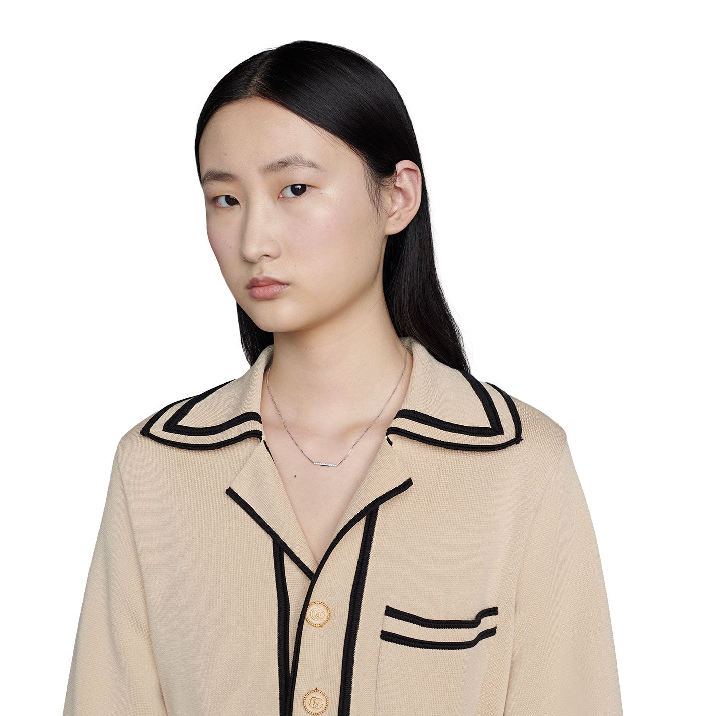 A person with long, dark hair is wearing a beige top with black trim and buttons. They are gazing at the camera with a neutral expression, elegantly highlighted by the Gucci Link to Love 18K White Gold Diamond Pavé Bar Necklace Pendant from GUCCI Fine Jewellery. The white background further emphasizes their graceful appearance.
