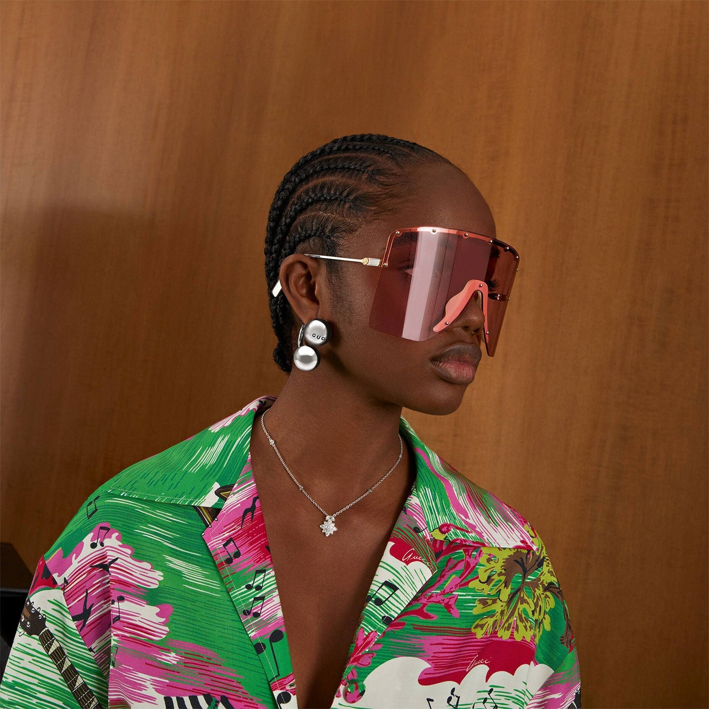 A person with braided hair wears oversized pink sunglasses and pearl earrings. Complementing their look is the Gucci Flora 18K White Gold Diamond Station Necklace Pendant from GUCCI Fine Jewellery. They wear a colorful patterned shirt featuring green and pink hues, reminiscent of Gucci Flora, set against a wooden backdrop.