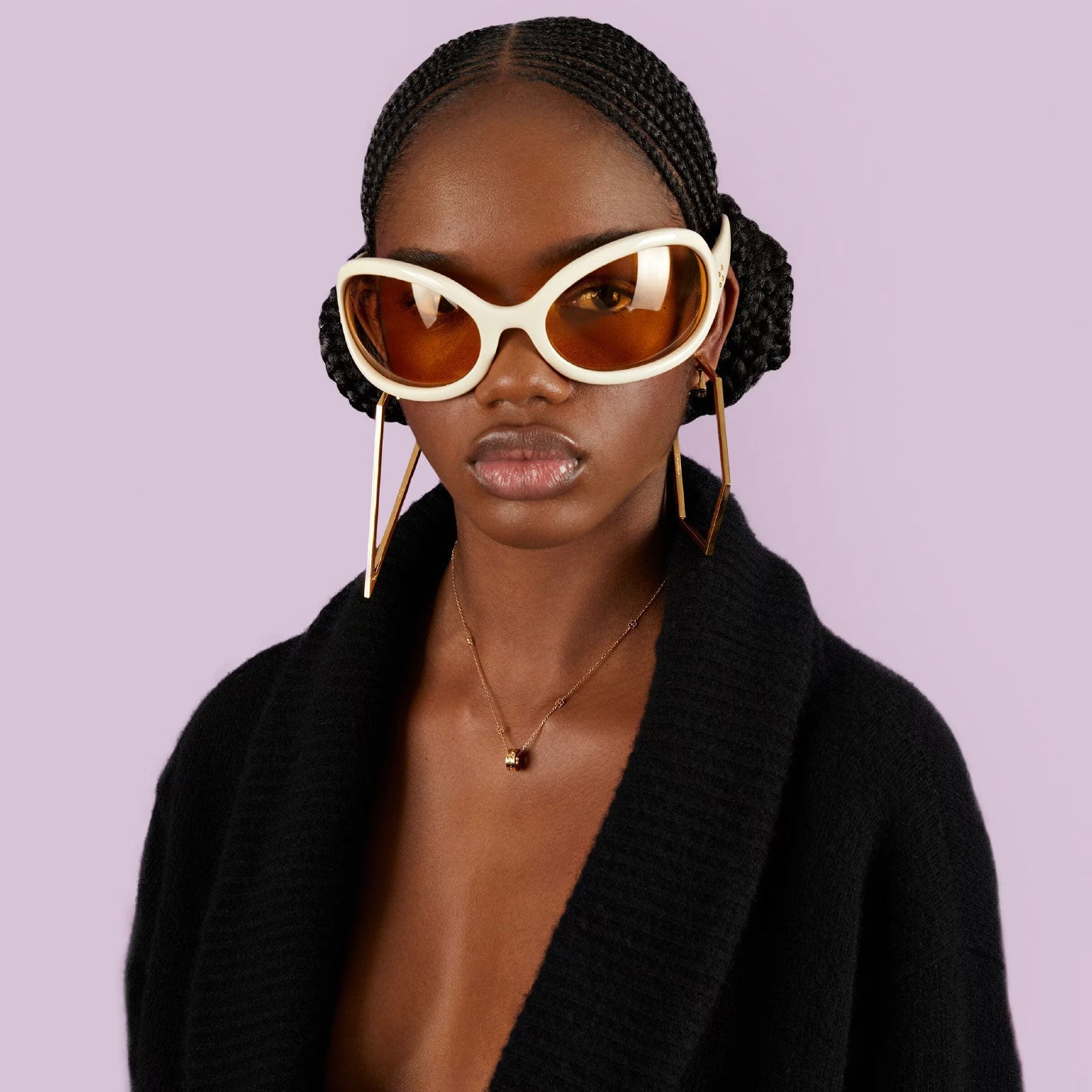 A person wearing oversized white sunglasses and elongated geometric gold earrings, with braided hair styled into two buns and adorned with a Gucci Icon Star 18K Yellow Gold Necklace Pendant by GUCCI Fine Jewellery. They wear a black outfit against a solid light purple background, which complements the look perfectly.