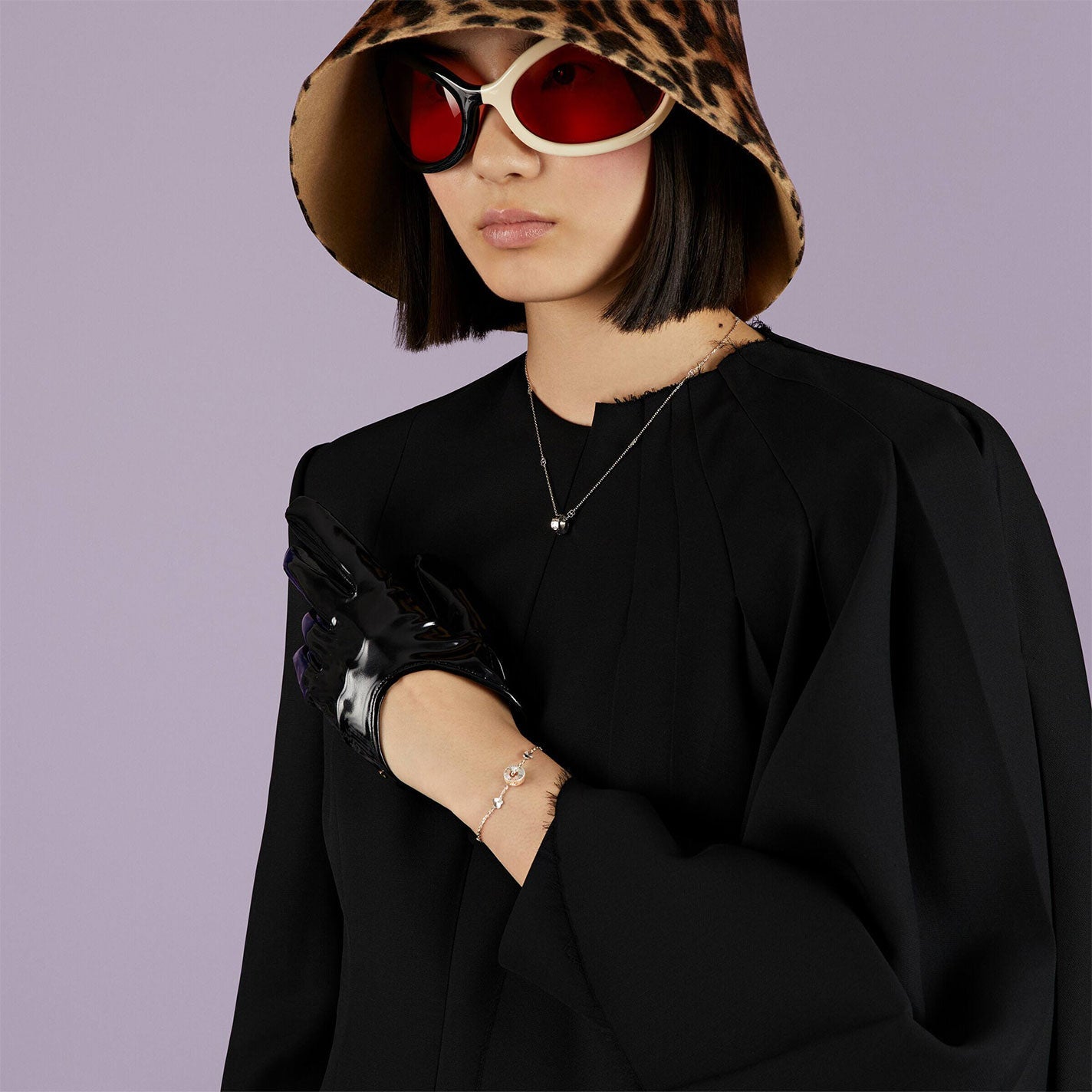 A person wearing a leopard print hat and large sunglasses poses against a purple background. They are dressed in a black outfit with a glove on one hand, accessorized with a Gucci Icon Heart 18K White Gold Necklace Pendant and bracelet from GUCCI Fine Jewellery.