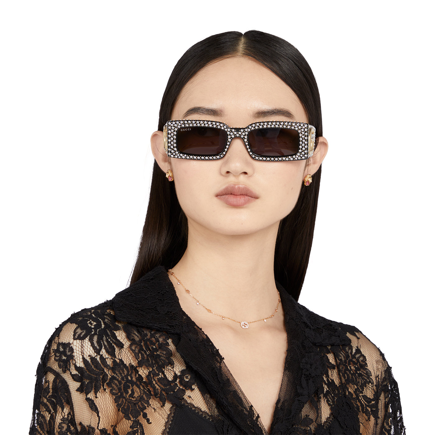 A woman stands against a plain background, wearing embellished rectangular sunglasses and a black lace top. Her straight black hair complements the sparkle of her diamond earrings and the Gucci Interlocking G 18K Rose Gold Necklace by GUCCI Fine Jewellery, while GG Pavé rings add an elegant touch to her ensemble.