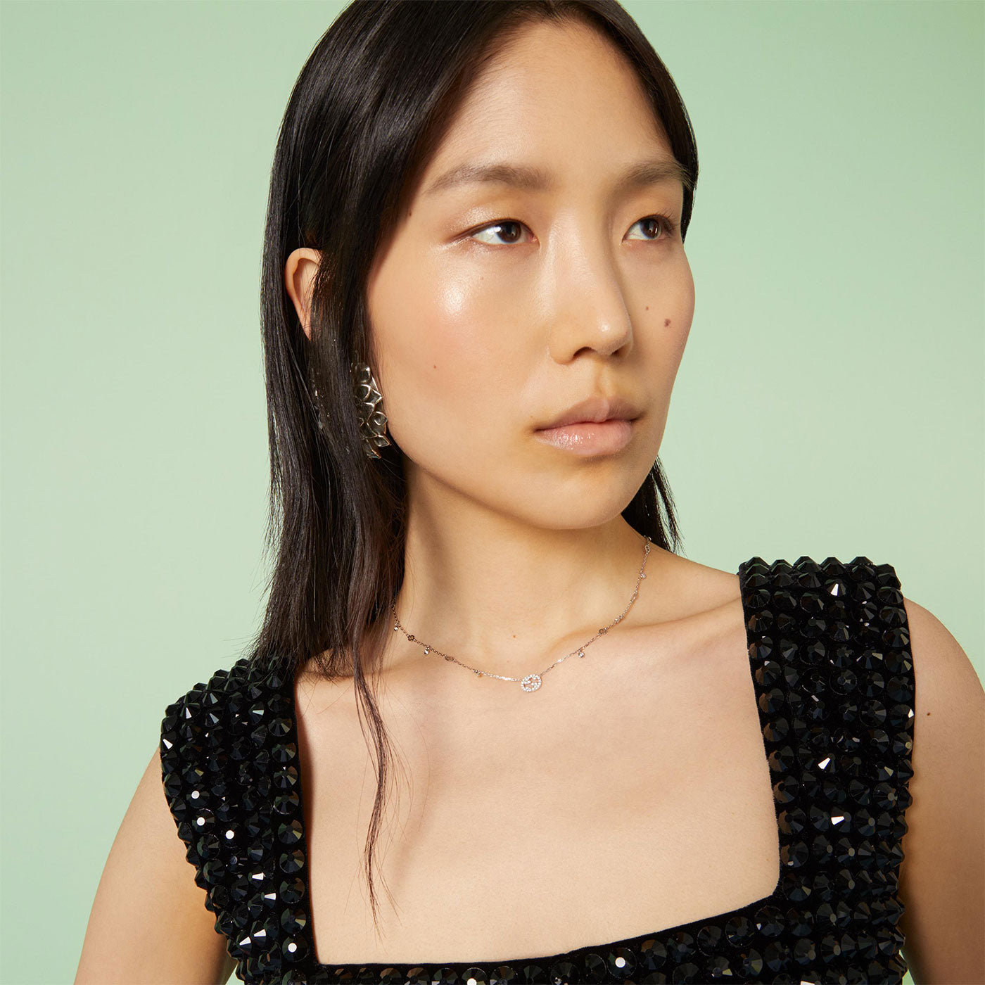 A woman with long hair wears a sleeveless black top adorned with sequins, her neck graced by the Gucci Interlocking G 18K White Gold Diamond Necklace from GUCCI Fine Jewellery. Her ornate earrings catch the light as she stands against a soft green background, gazing to the side.