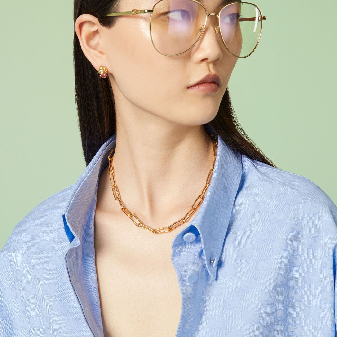 A person wearing a light blue button-up shirt with a subtle pattern, large round glasses, and gold earrings with pink accents stands against a pale green background, elegantly accessorized by the Gucci Link to Love 18K Yellow Gold Wide Chain Necklace from GUCCI Fine Jewellery.