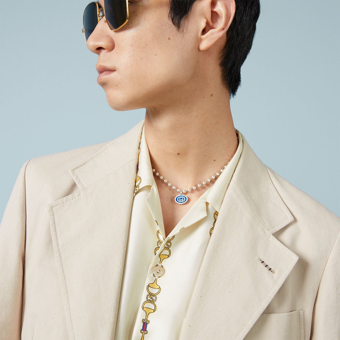 A person in a light beige blazer and patterned shirt looks to the side, sporting sunglasses and a Gucci Interlocking G Sterling Silver Boule Chain Necklace Pendant from GUCCI Fine Jewellery. The background is plain light blue.