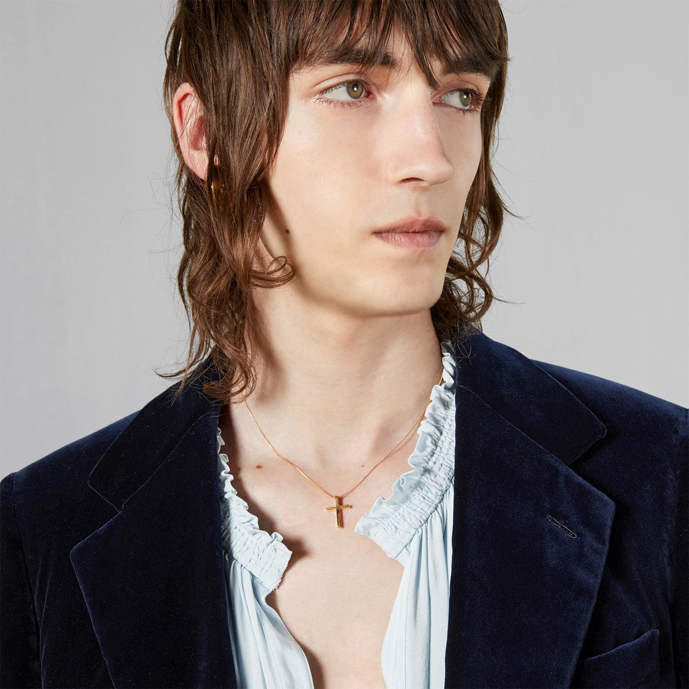 A person with shoulder-length brown hair wears a dark blue velvet blazer and a frilled light blue shirt. They accessorize with the GUCCI Fine Jewellery Link to Love Cross 18K Yellow Gold Necklace Pendant, gazing off to the side against a neutral background.