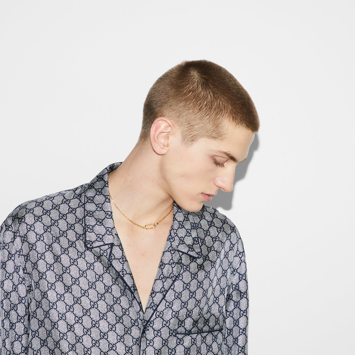 A person with short hair, in a patterned button-up shirt and a GUCCI Fine Jewellery Link To Love 18K Yellow Gold Chain Necklace, stands against a plain white background, looking down and to the side.