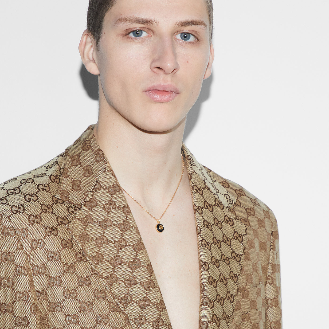 A person in a Gucci-patterned suit jacket shows off the GUCCI Fine Jewellery's Gucci Interlocking 18K Yellow Gold Chain Necklace with Onyx Pendant, featuring the subtle Interlocking G logo, all set against a plain white background.