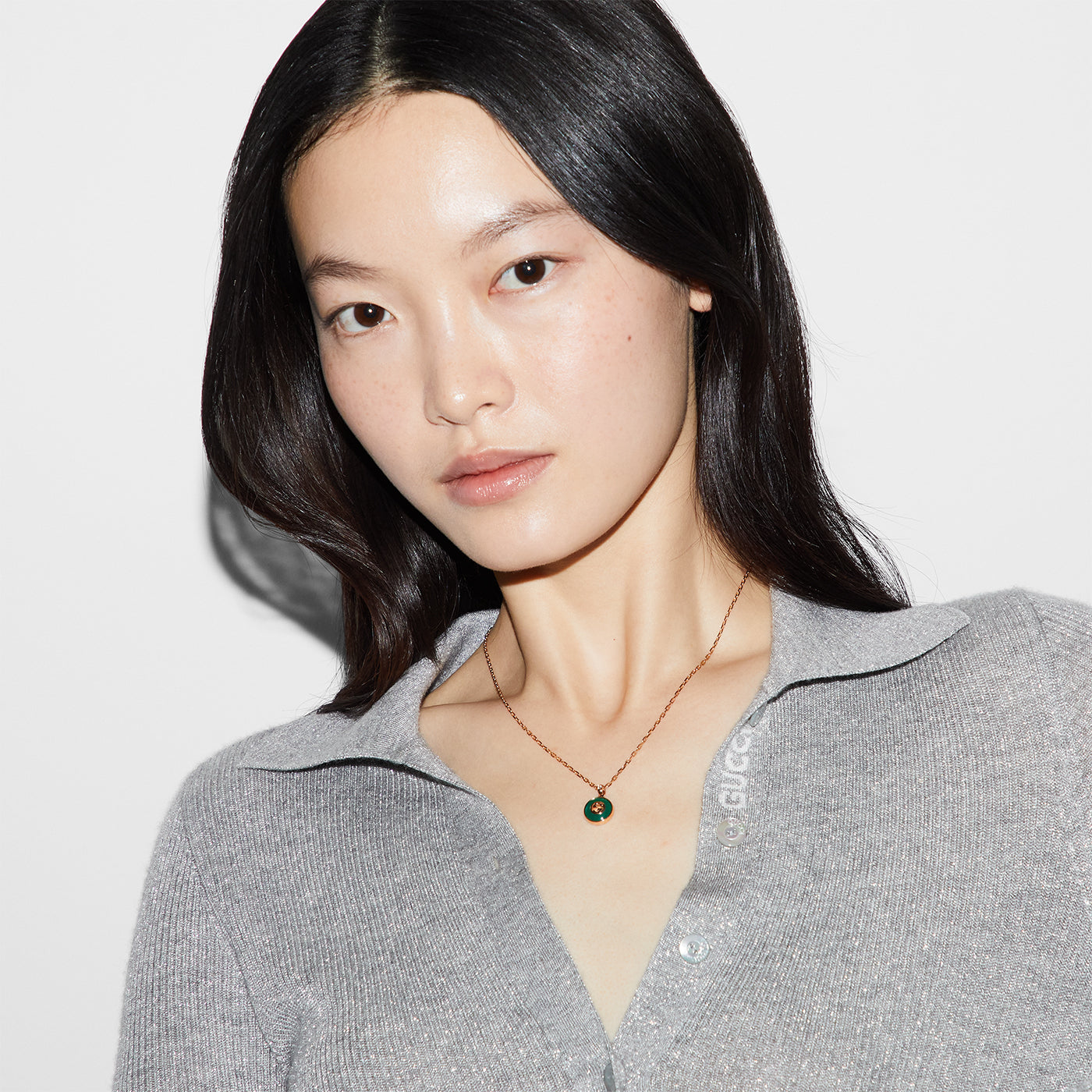A person with long dark hair is wearing a gray top and an adjustable Gucci Interlocking 18K Rose Gold Chain Necklace featuring a striking circular green agate pendant. They pose against a plain white background, highlighting the GUCCI Fine Jewellery piece.