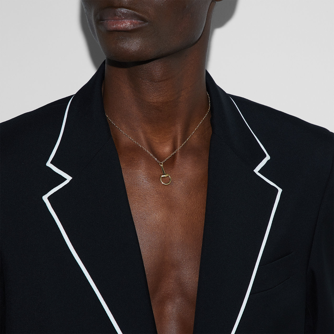 A person in a black blazer with white piping highlights their bare chest with a GUCCI Fine Jewellery Horsebit 18K Yellow Gold Chain Necklace against a plain white wall.