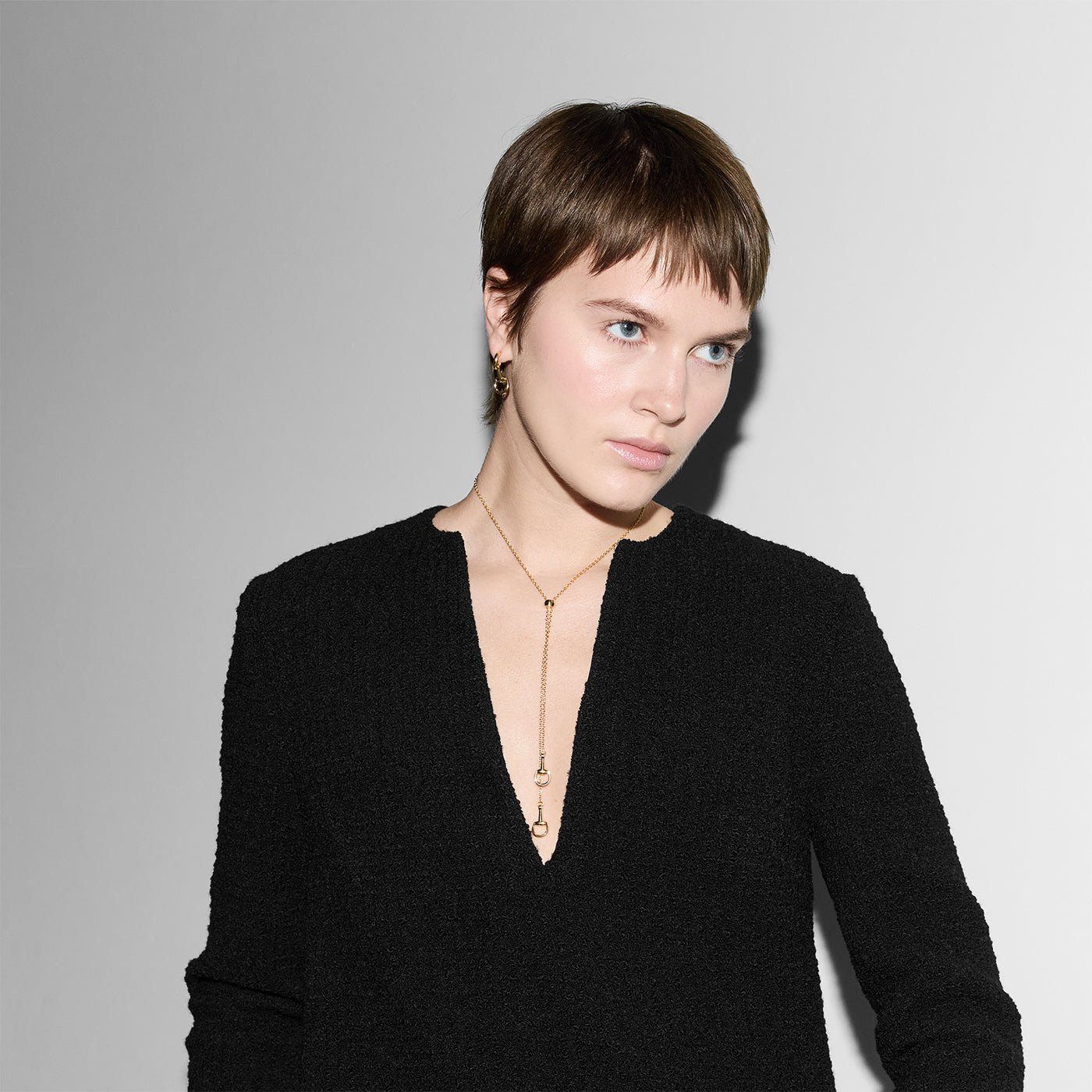 A person with short hair in a sleek black outfit, wearing a Gucci Horsebit 18K Yellow Gold Pendant Necklace by GUCCI Fine Jewellery, looks slightly aside. The necklace's subtle elegance complements the white and gray background.