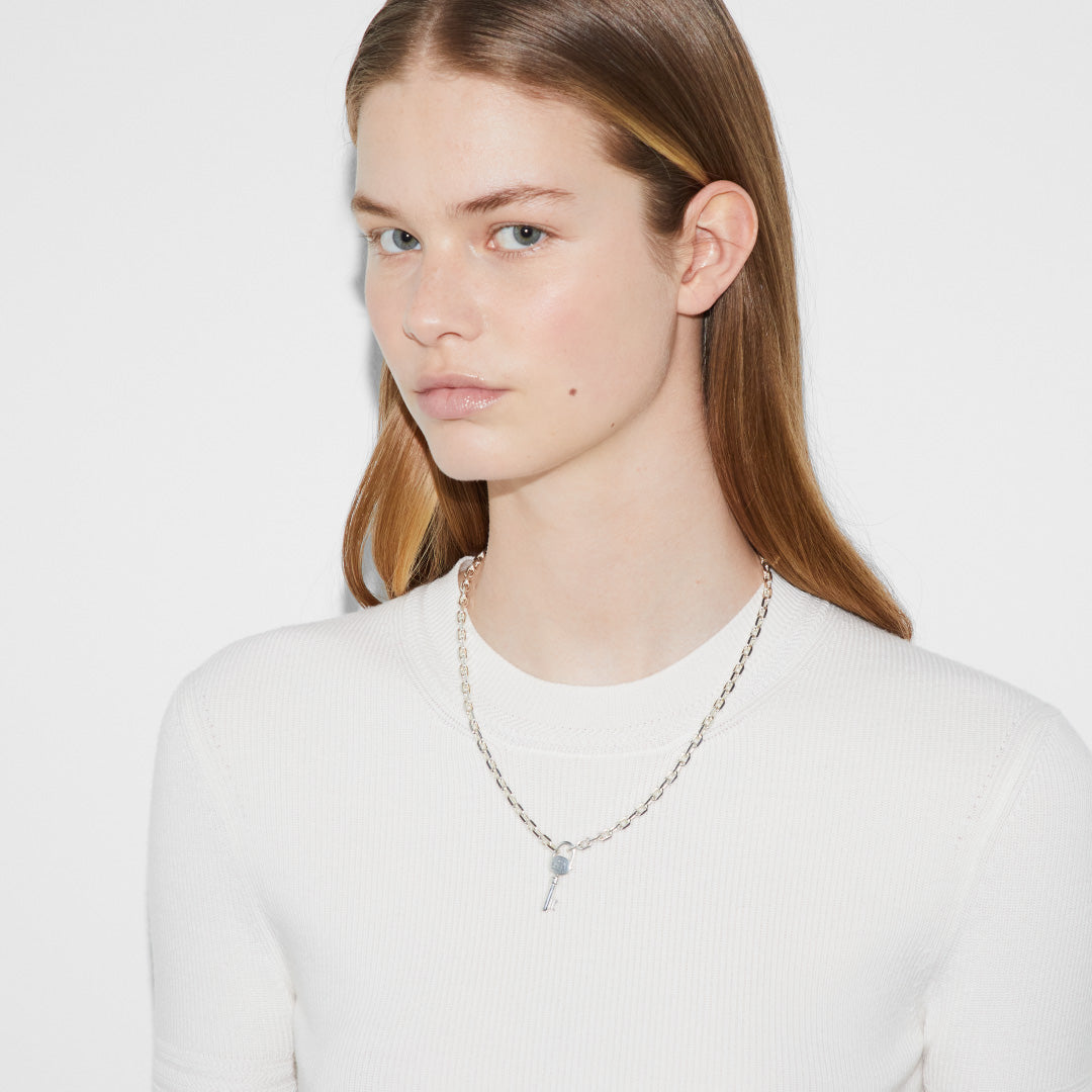 A person with long, straight hair is wearing a white ribbed shirt and a Gucci Trademark Sterling Silver Necklace Pendant by GUCCI Fine Jewellery. The background is plain and white.