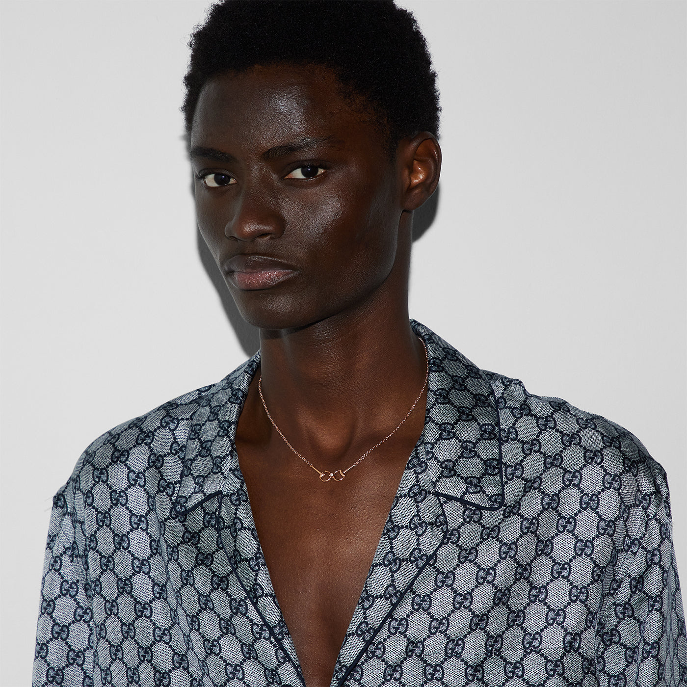 A person wears an 18K rose gold necklace from GUCCI Fine Jewellery. The shirt, with a geometric diamond-like pattern, complements the Gucci Horsebit Diamond Chain Necklace. They maintain a neutral expression against a plain backdrop.