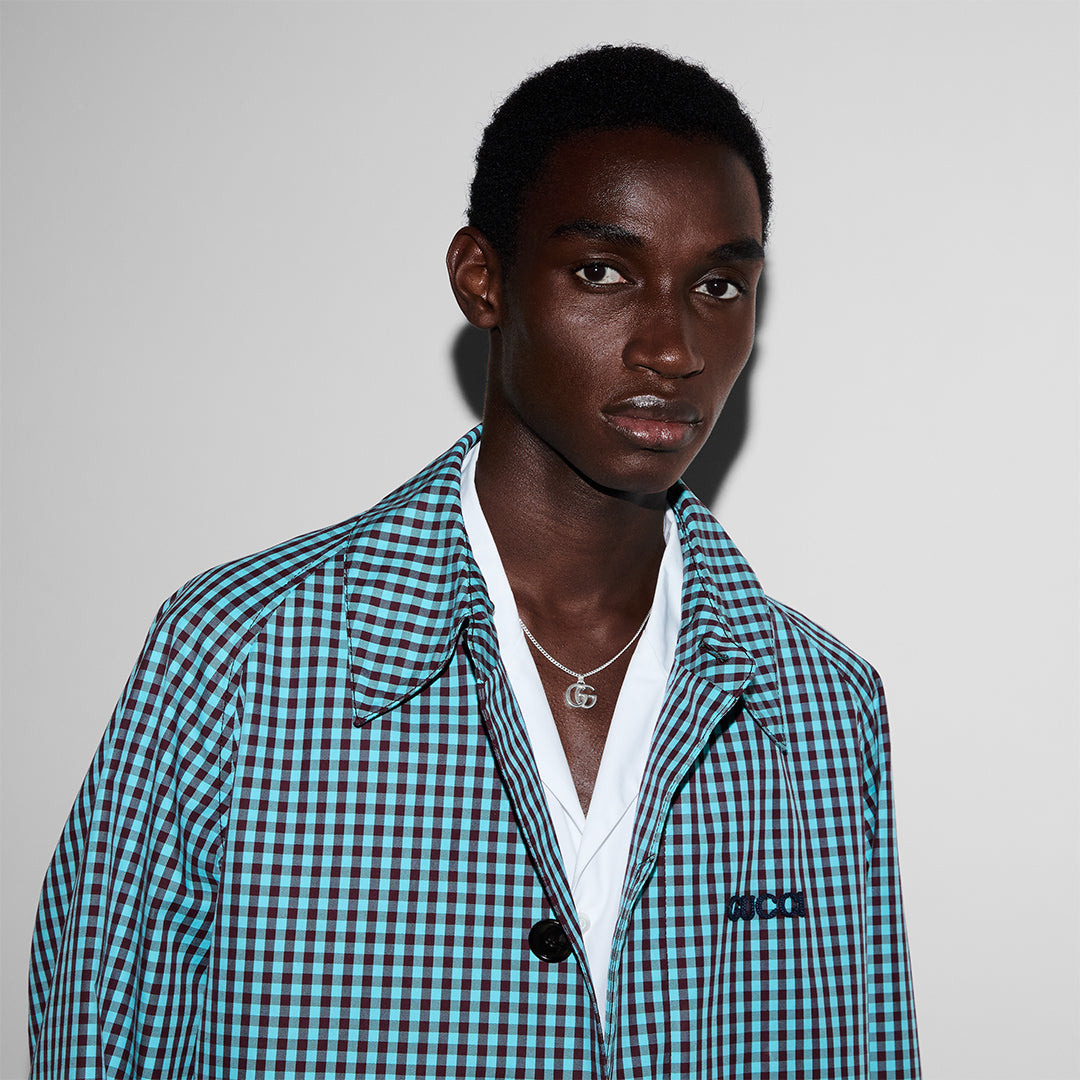 A young man wearing a blue and black checkered jacket over a white shirt, accessorized with a Gucci GG Marmont Sterling Silver Necklace Pendant from GUCCI Fine Jewellery, stands against a plain light gray background.