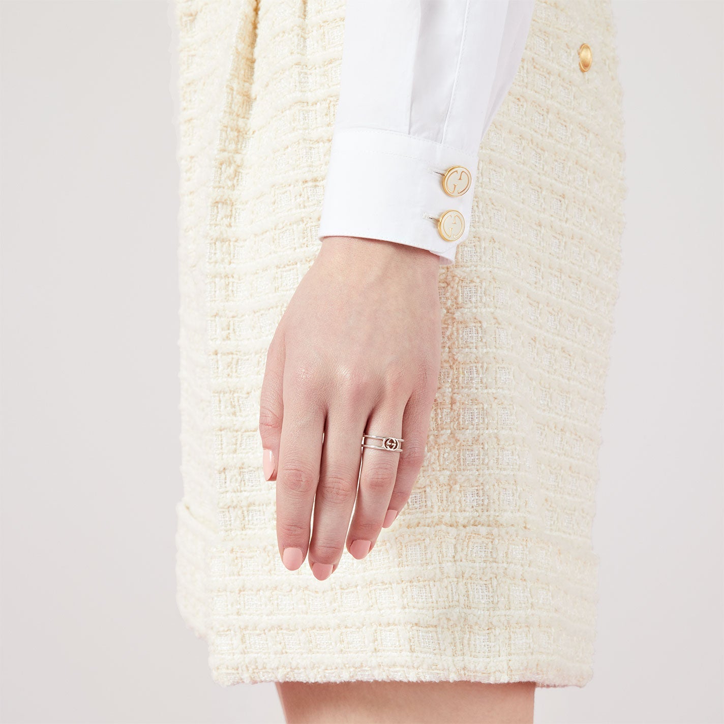 A close-up of a person's hand flaunting the Gucci Interlocking G Thin Open Band Sterling Silver Ring by GUCCI Fine Jewellery, elegantly engraved. The person is dressed in a textured cream skirt and a white blouse with gold buttons on the cuff. The background is a neutral, light shade, perfectly highlighting the allure of the Interlocking G design.