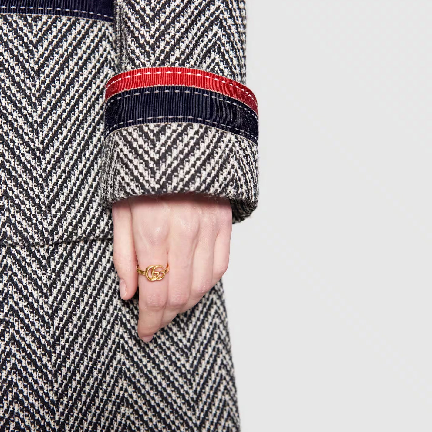 A close-up captures an individual wearing a herringbone-patterned coat, accentuated by red and navy stripes on the sleeve, as they display the GUCCI Fine Jewellery's Gucci Running G 18K Yellow Gold Ring. Set against a plain light-colored background, this elegant accessory, crafted in Italy, is prominently highlighted.